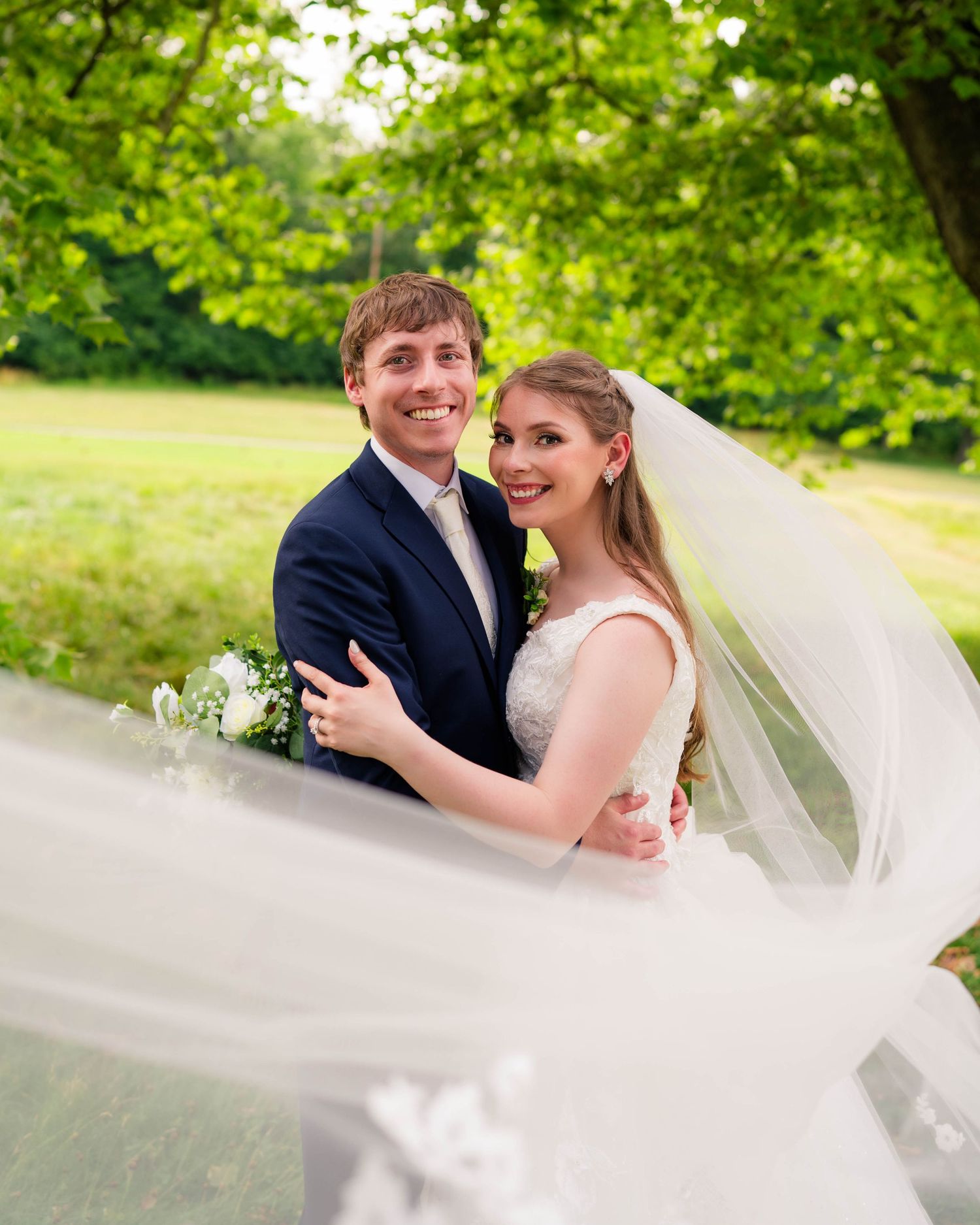 Smiling bride and groom embrace outdoors as flowing cathedral veil billows in the breeze among lush green trees.