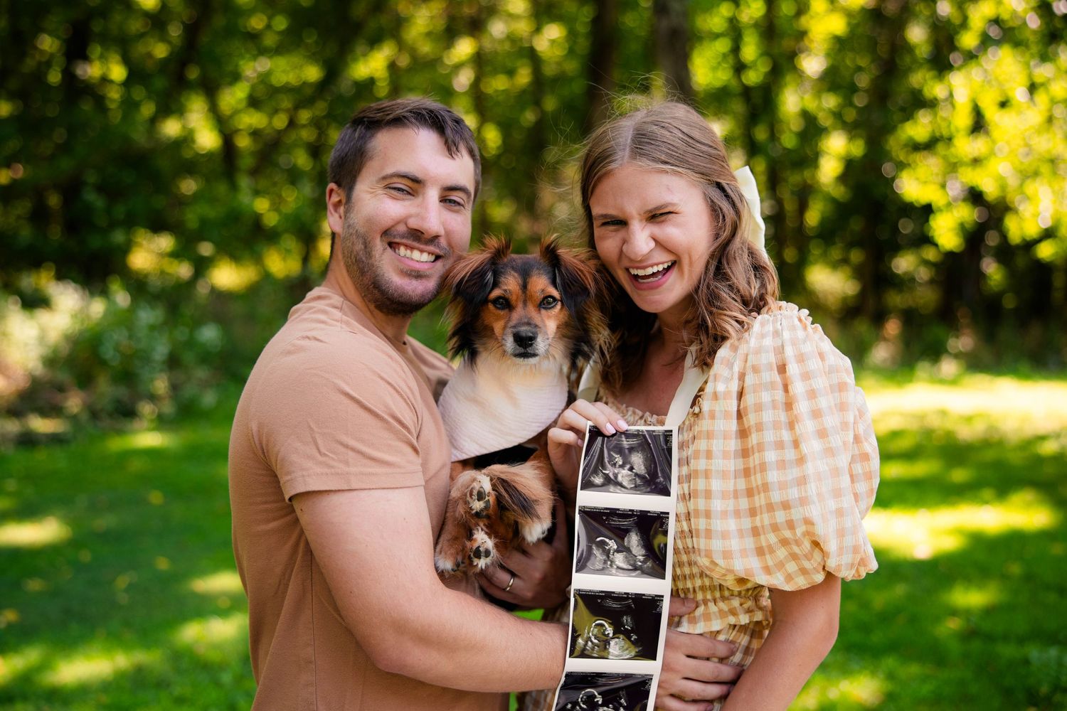 Laughing couple holding dog and ultrasound photos outdoors in lush green park setting.