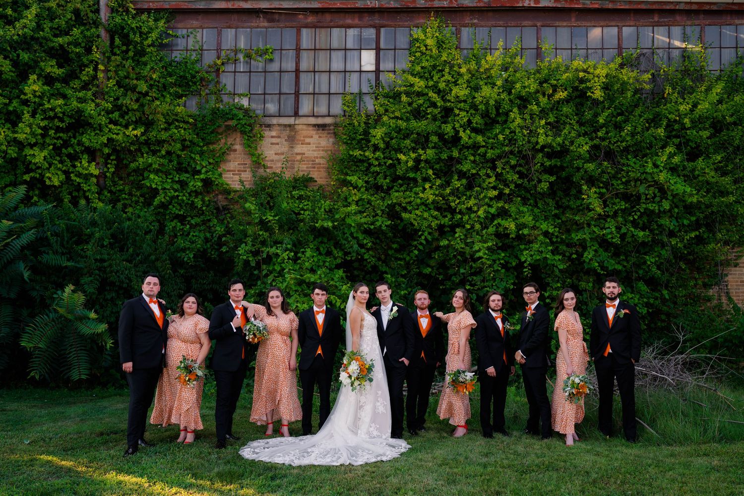 Wedding party in orange bridesmaid dresses and black suits posing before lush green industrial backdrop.