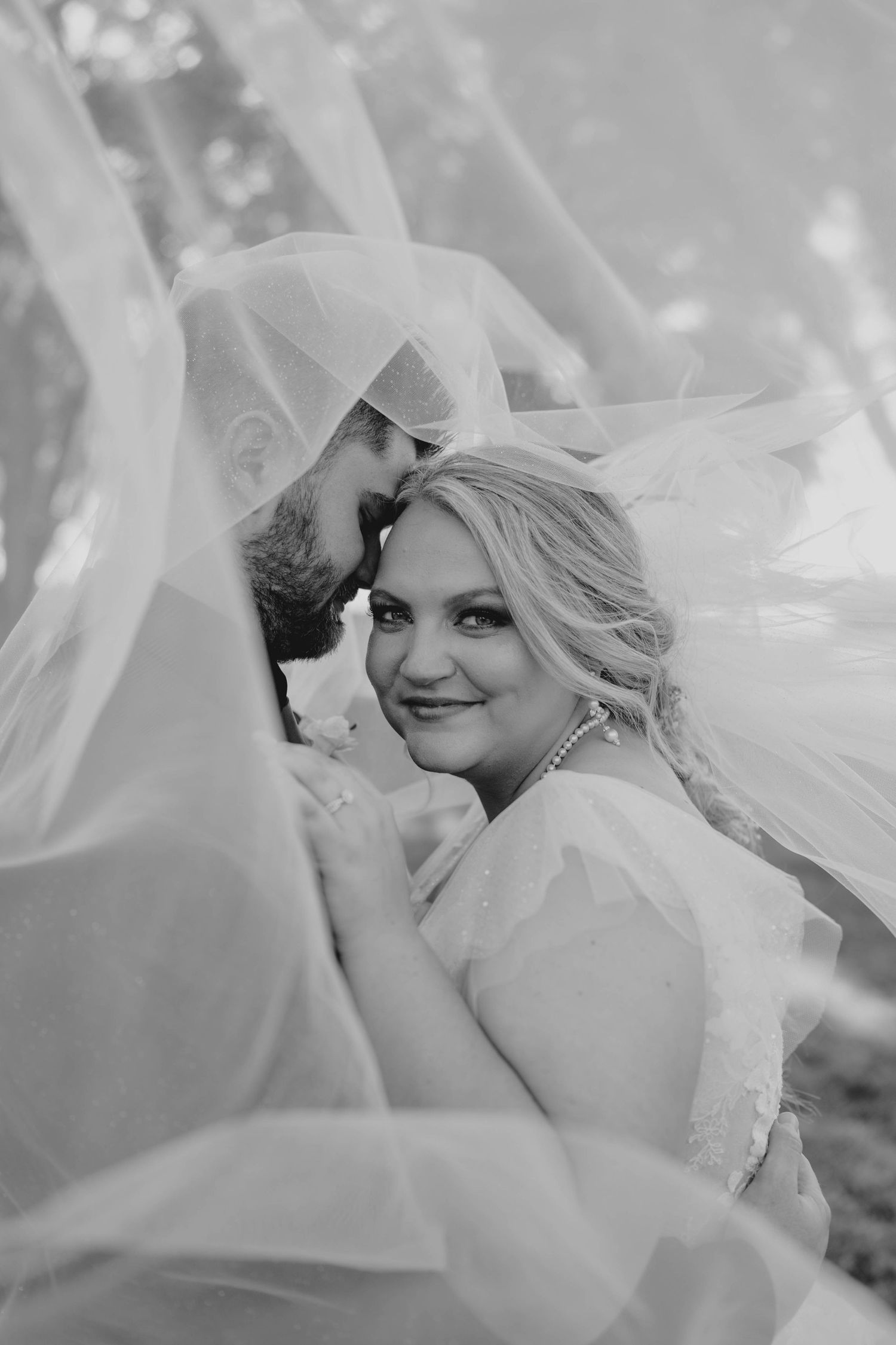 Black and white wedding photo of bride and groom embracing under flowing veil, bride smiling at camera.