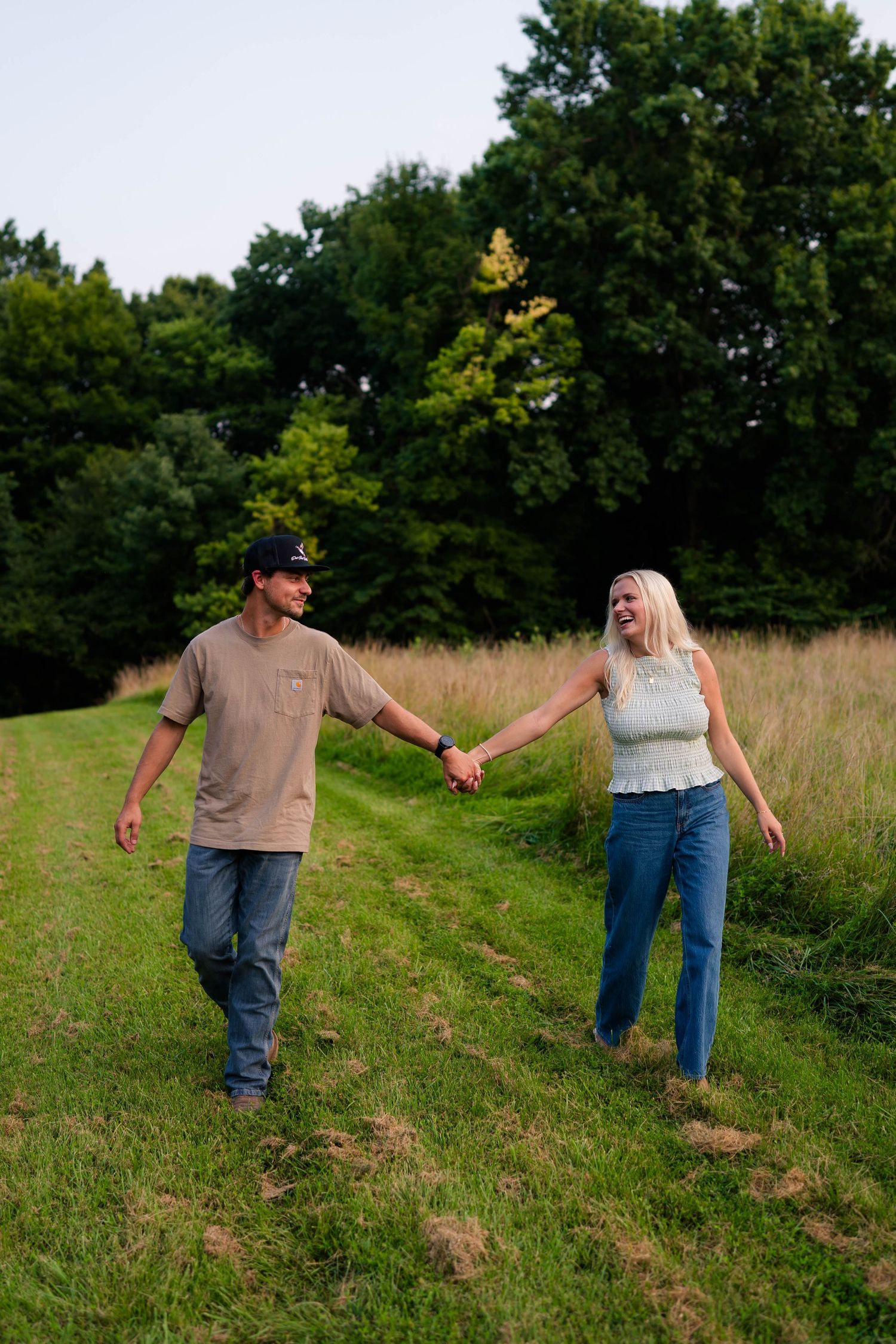 A couple holds hands and laughs while walking along a green grass path surrounded by tall golden fields and trees.