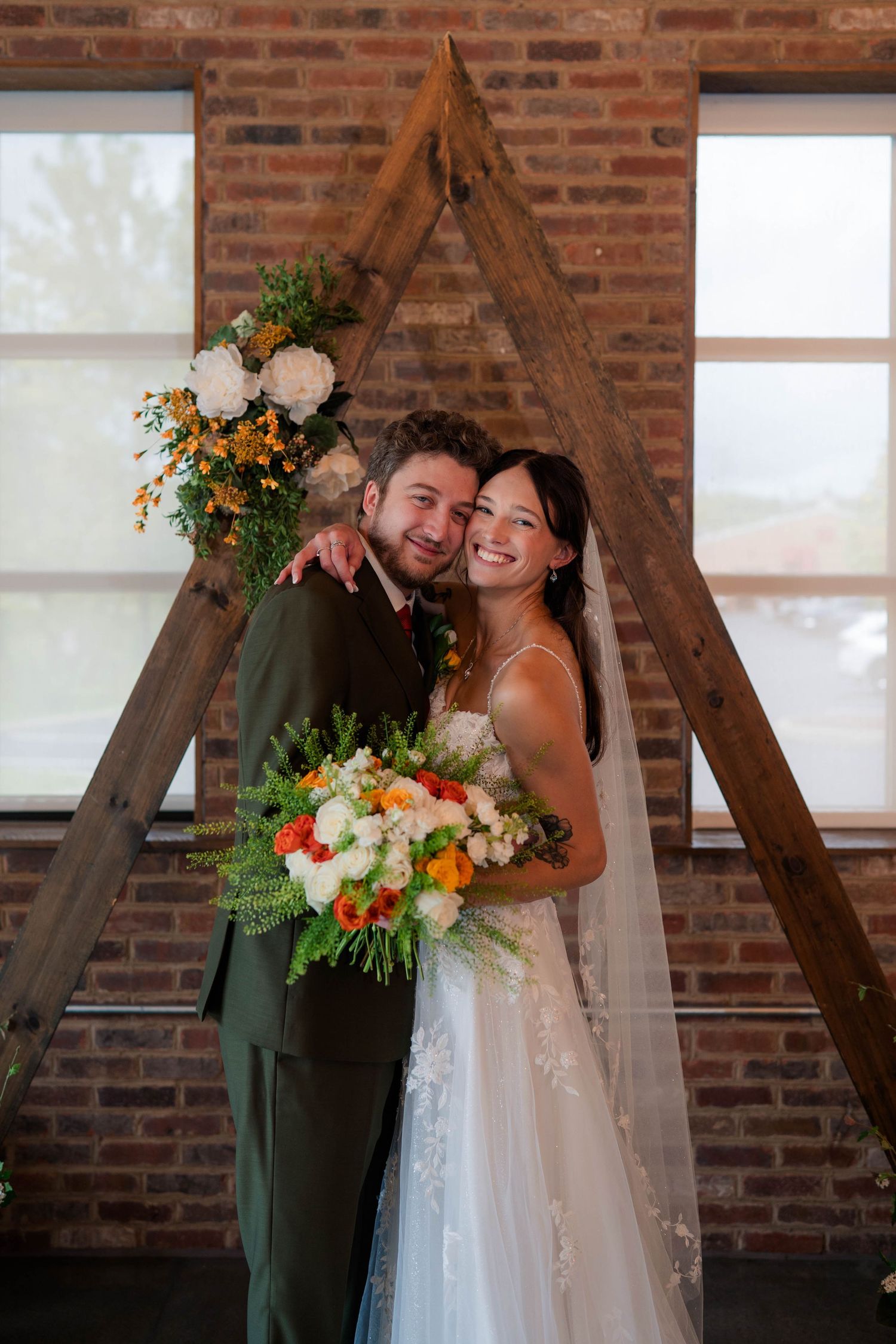 Happy newlyweds embracing under rustic wooden triangular arch with floral arrangements at reception.