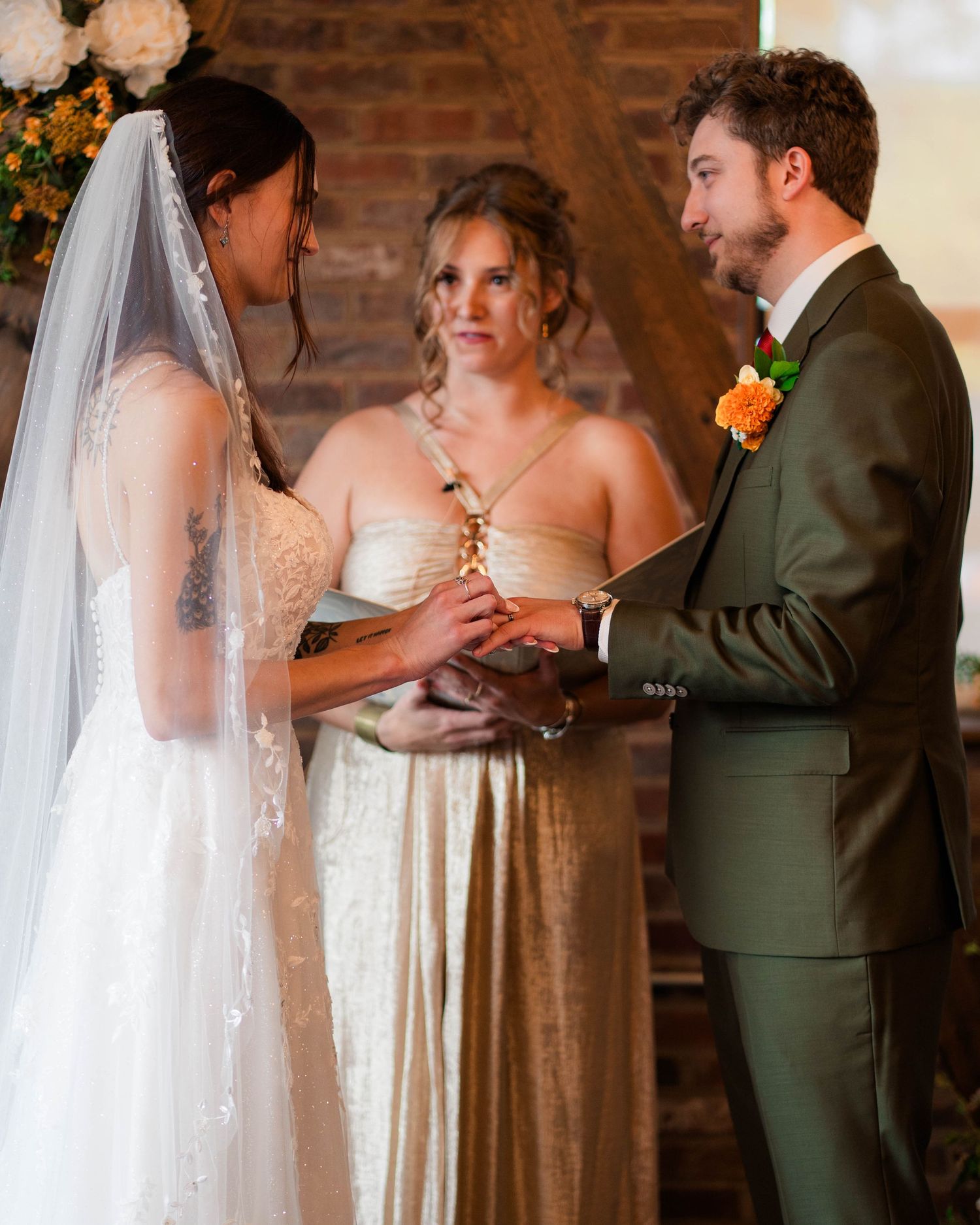 Bride and groom exchange vows at wedding ceremony with officiant in golden dress at rustic brick venue.
