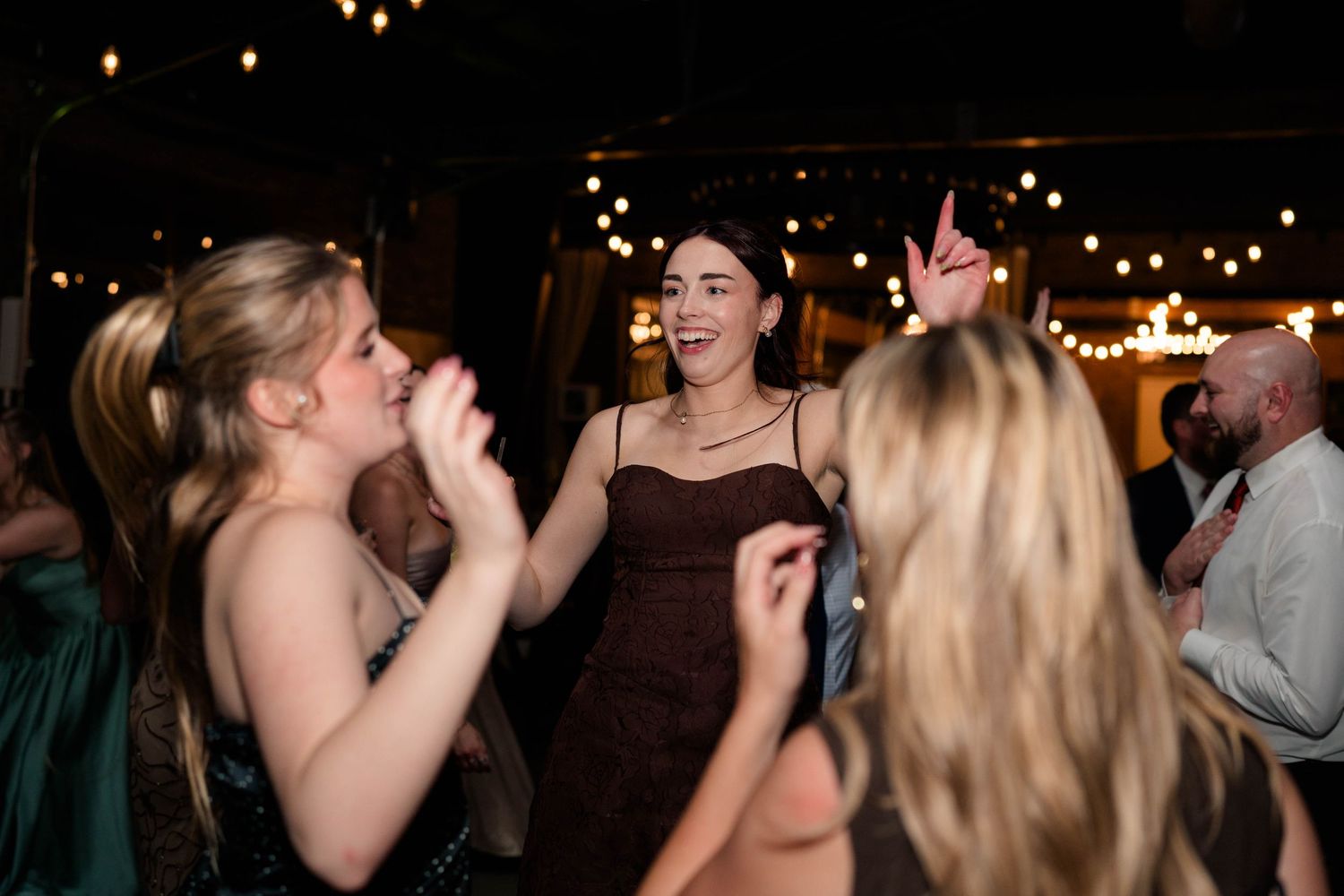 Women in formal attire dancing and celebrating joyfully under string lights at wedding reception.