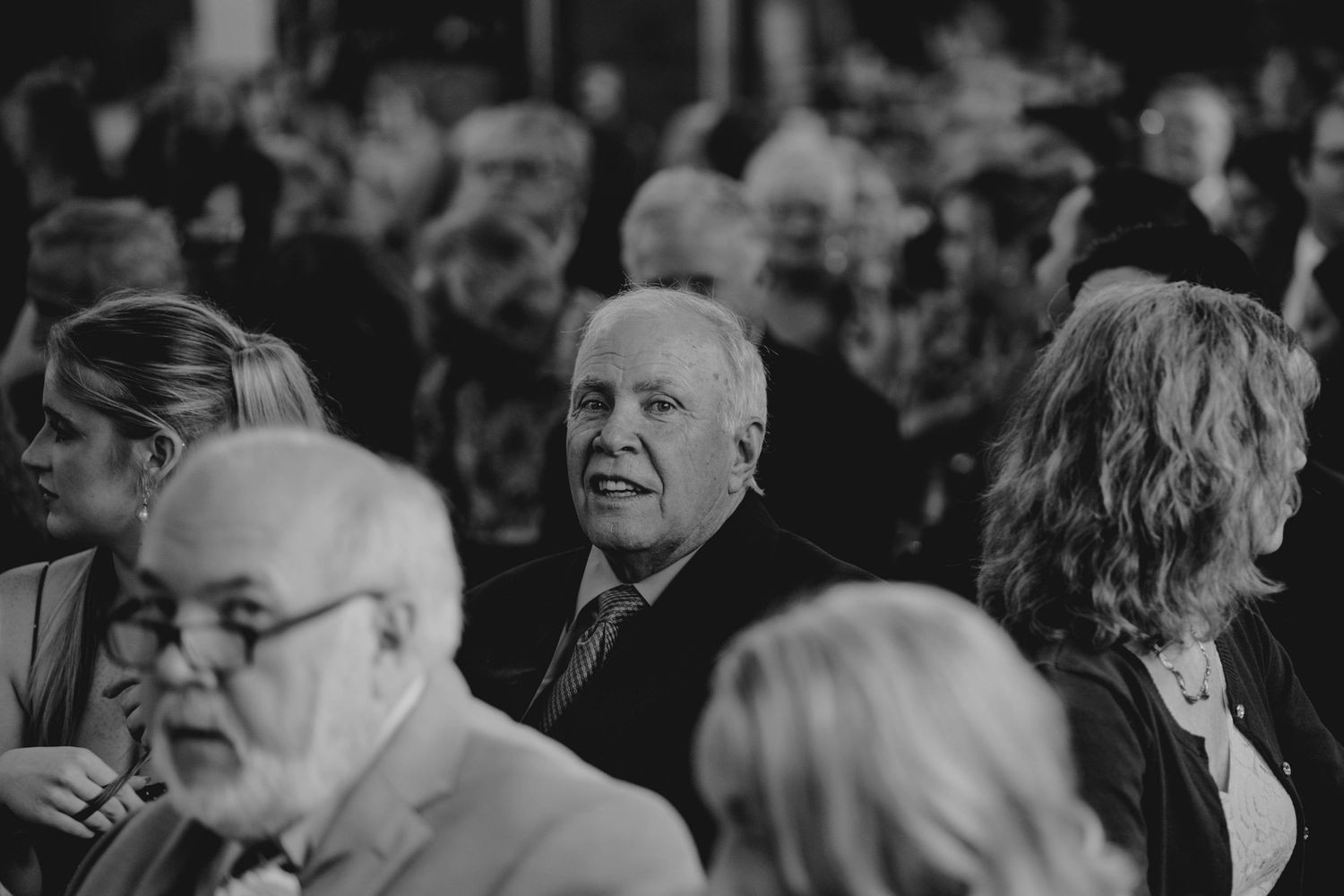 Black and white candid photo of wedding guests seated and watching ceremony in crowded venue.