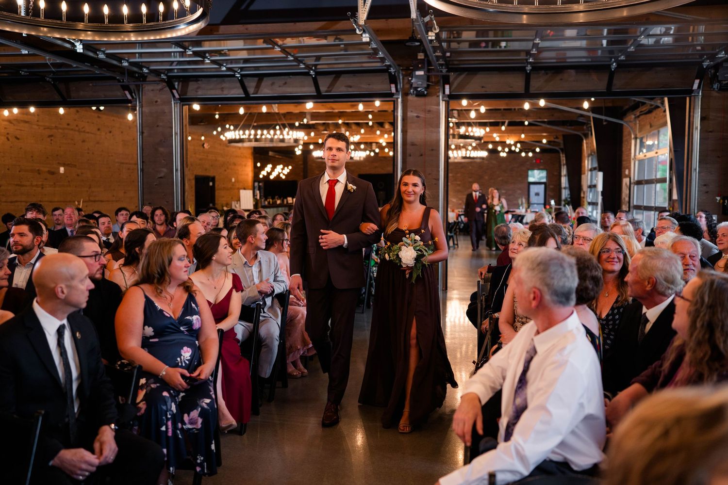 Wedding party processional with groomsman and bridesmaid walking down the aisle in a brick-walled reception hall.