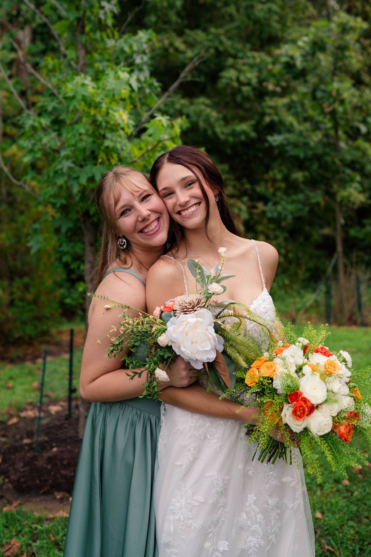 Bride in lace gown embraces smiling bridesmaid in sage green dress while holding colorful wedding bouquet outdoors.