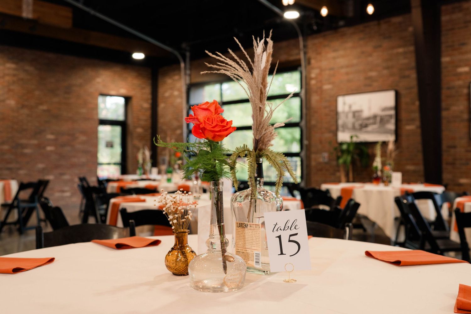 Elegant wedding reception table centerpiece featuring an orange rose and dried grasses in glass vases with table number.