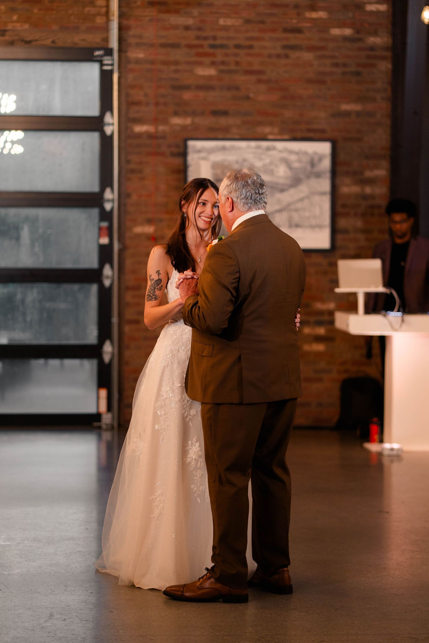 Bride in lace gown sharing a father-daughter dance in a rustic brick-walled wedding reception venue.