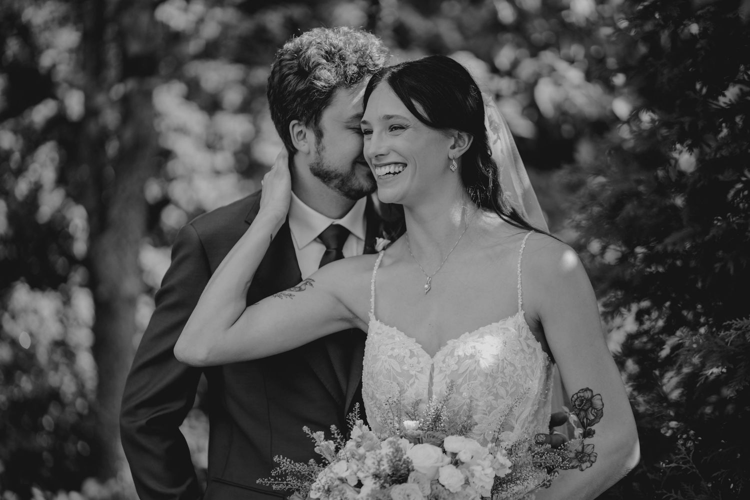 Black and white photo of groom whispering to smiling bride holding bouquet outdoors.