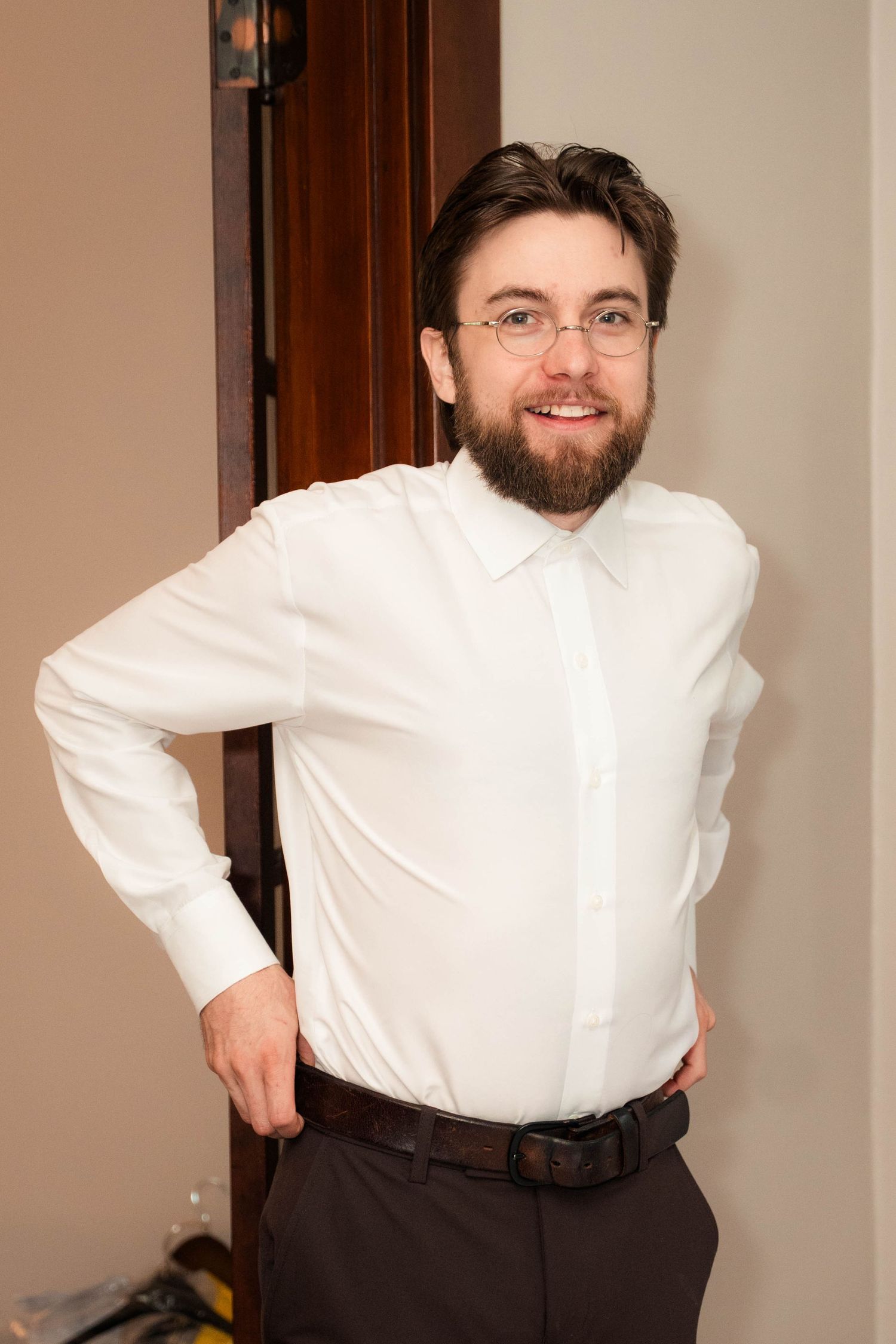 Smiling bearded man in white dress shirt and glasses posing confidently near a wooden door.