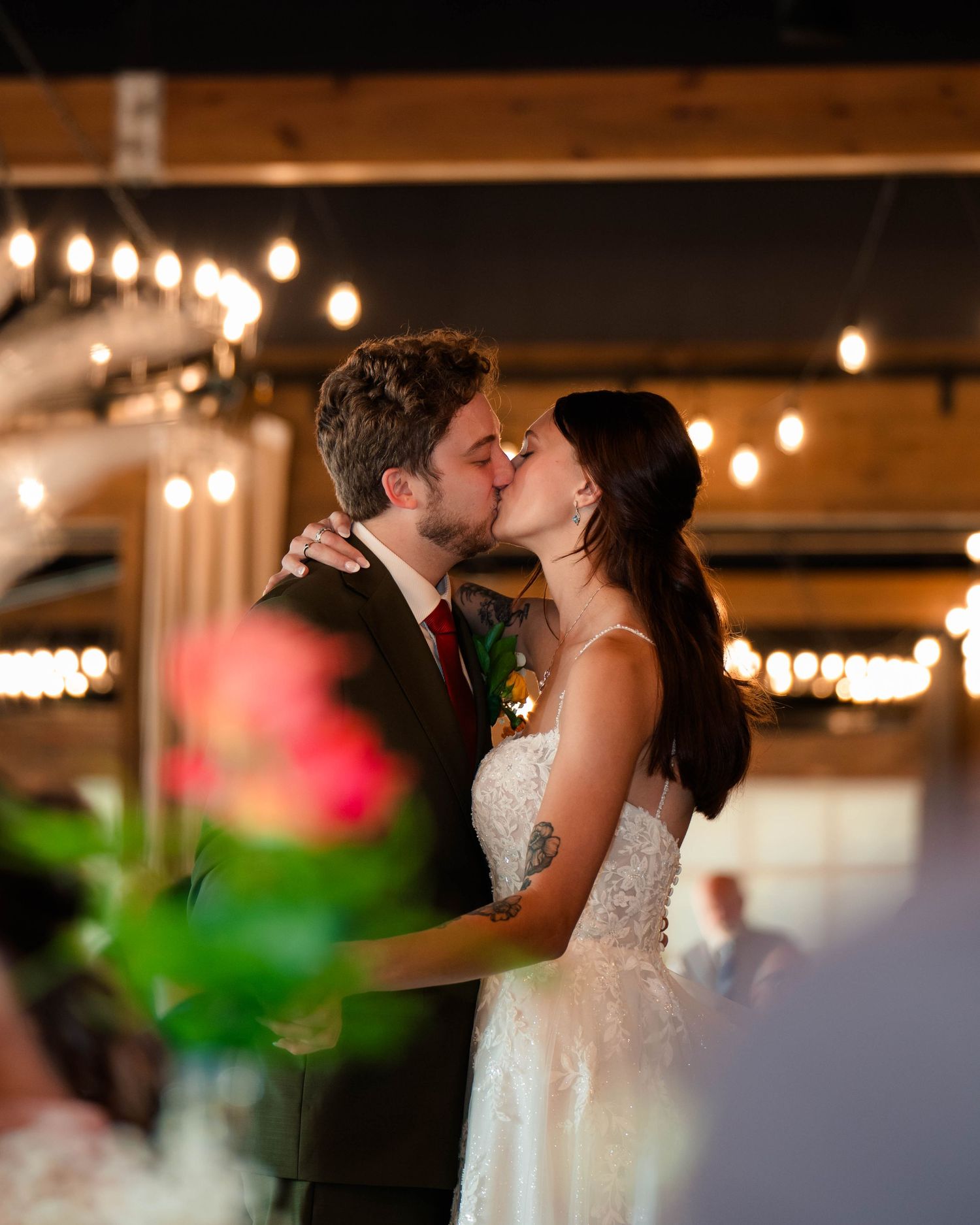 Bride and groom share a romantic first kiss under warm string lights at wedding reception.