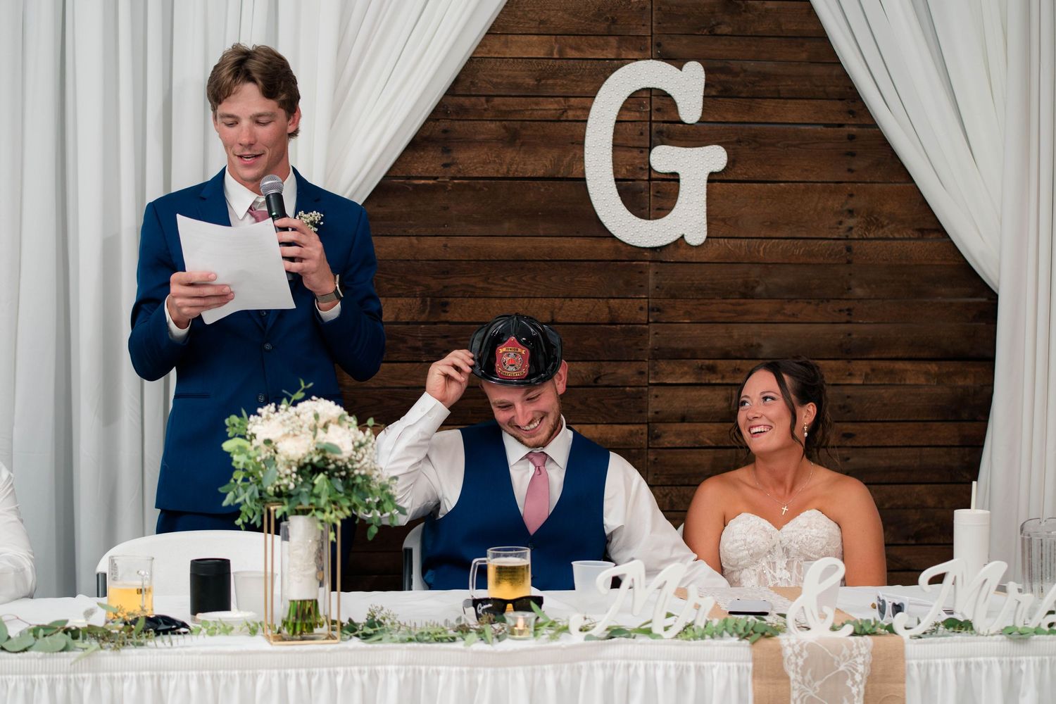 Best man delivers a heartfelt wedding speech at the head table while the bride and groom smile during the reception.