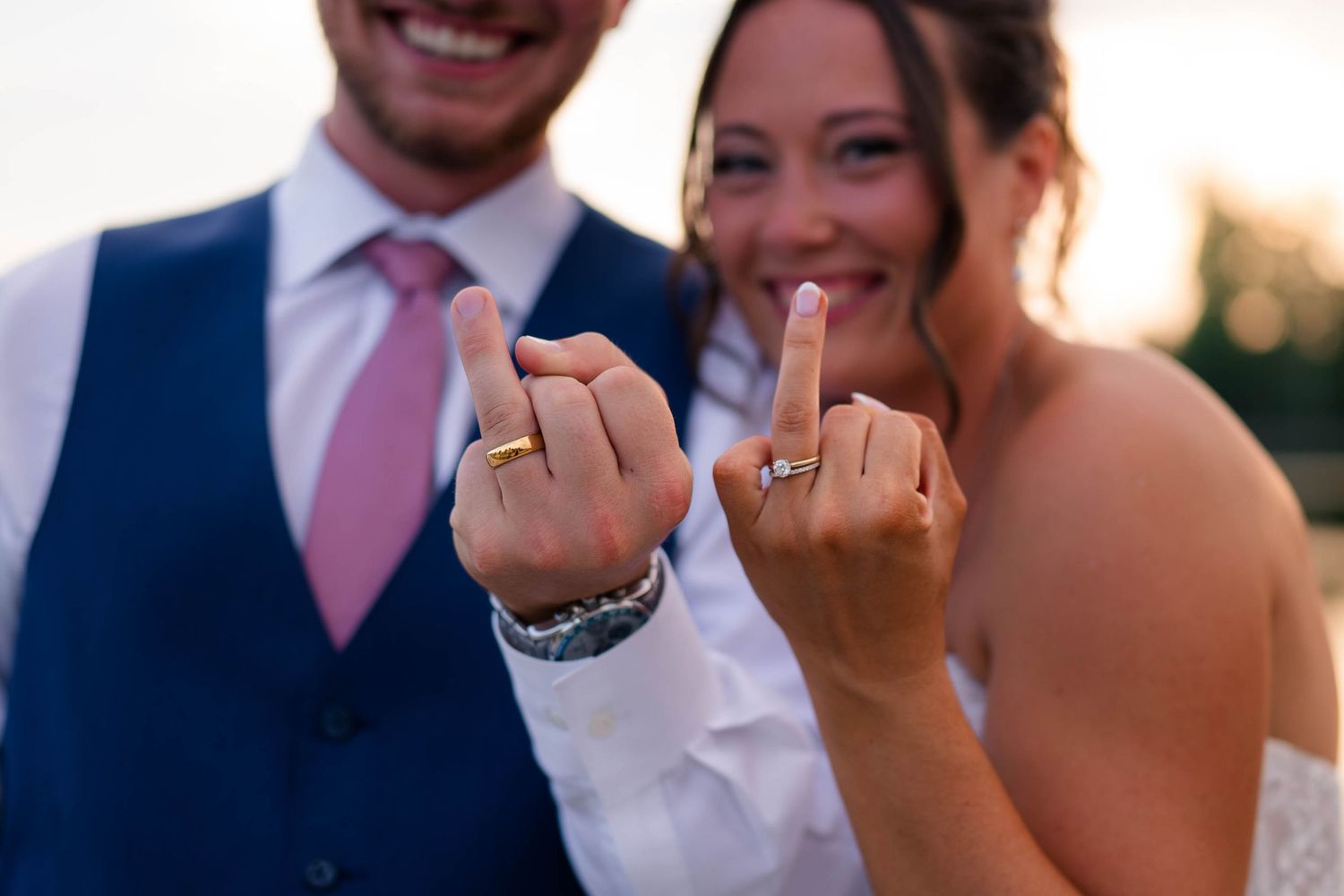 Smiling bride and groom showing off wedding rings on fingers at outdoor ceremony in golden hour light.