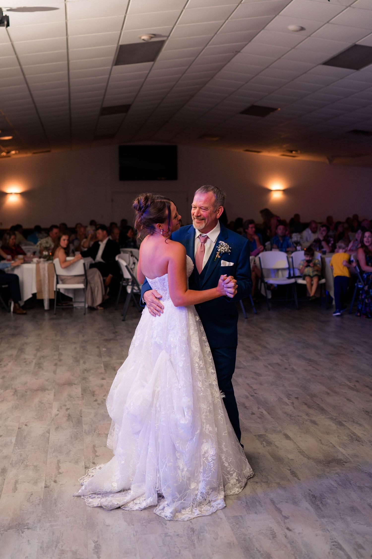 Bride shares a tender father-daughter dance on the wedding reception floor surrounded by seated guests.