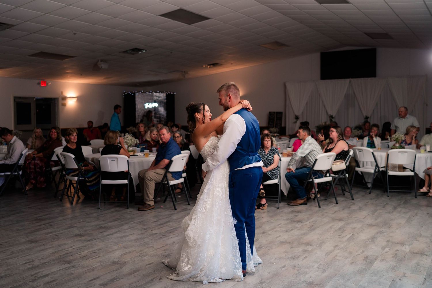 Bride and groom share romantic first dance surrounded by seated wedding guests in decorated reception hall.
