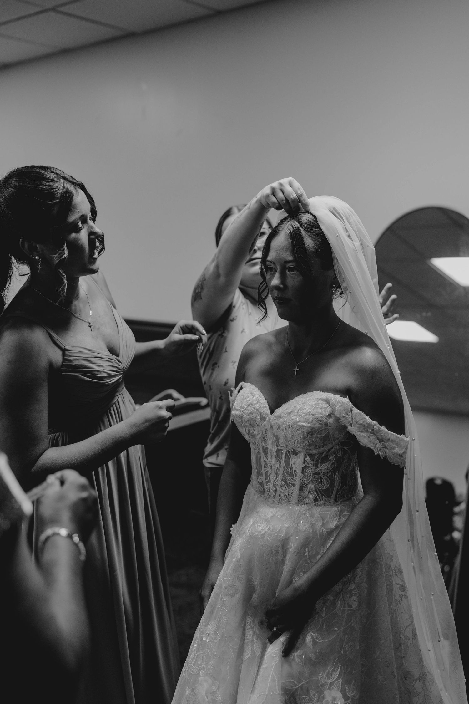 Black and white photo of bridesmaids helping a bride in lace gown put on her veil before the wedding ceremony.