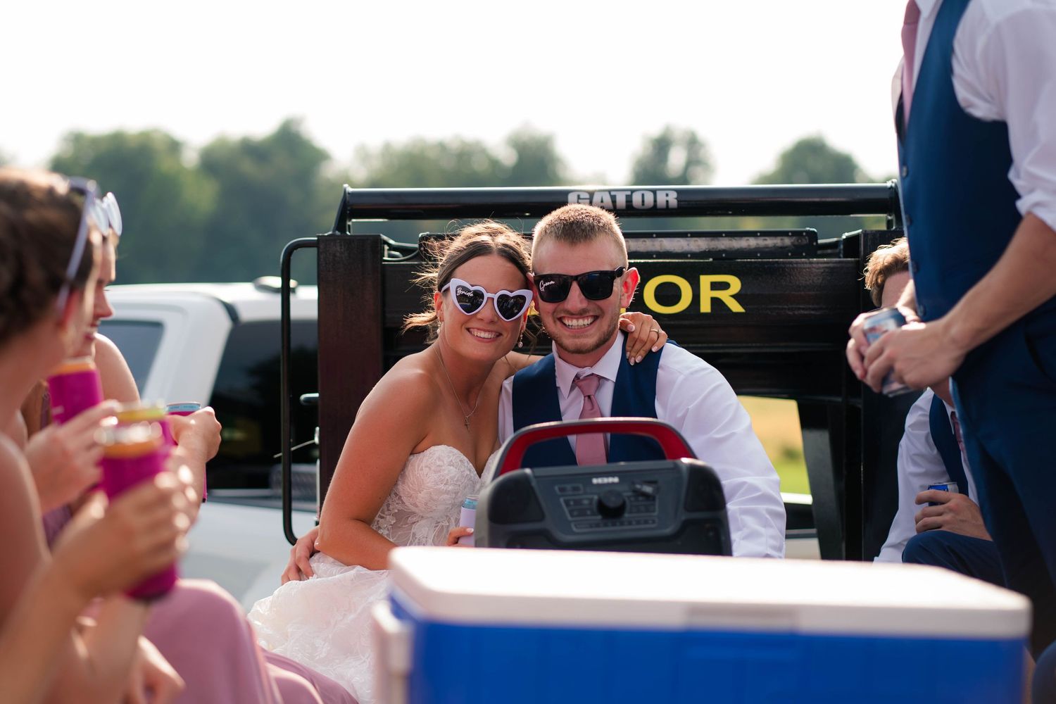 Smiling bride and groom wearing heart-shaped sunglasses riding in Gator utility vehicle at wedding.