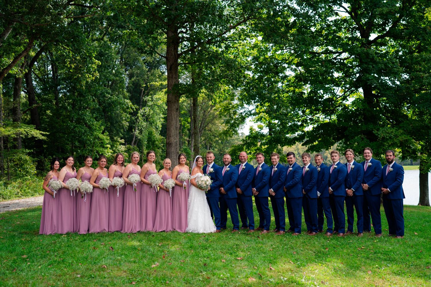 Wedding party poses outdoors, bridesmaids in mauve gowns and groomsmen in navy suits by a pond.