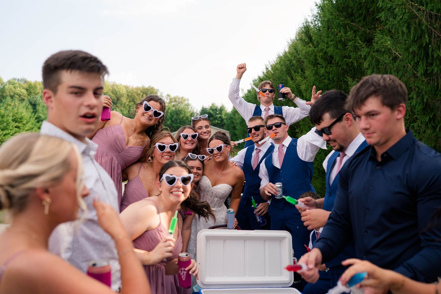 Wedding guests in sunglasses and formal attire pose and celebrate energetically outdoors at a summer wedding reception.