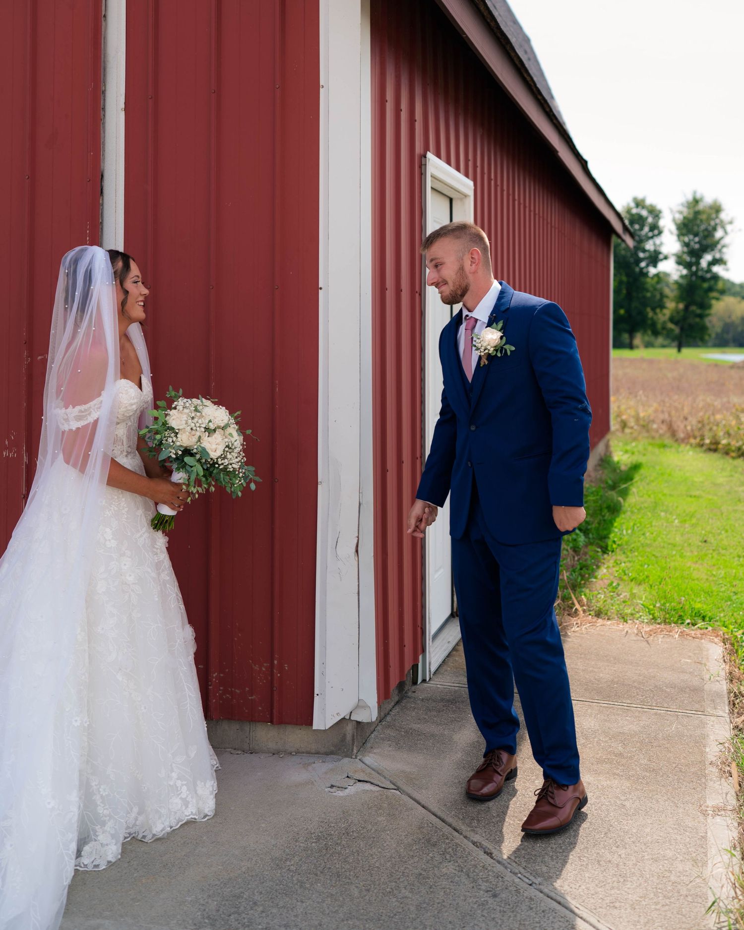 Bride and groom share first look moment outside red barn, bride holding white floral bouquet.