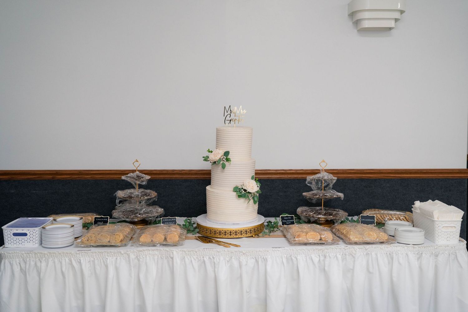 Elegant three-tier white wedding cake with greenery on a gold stand surrounded by desserts on a draped table.