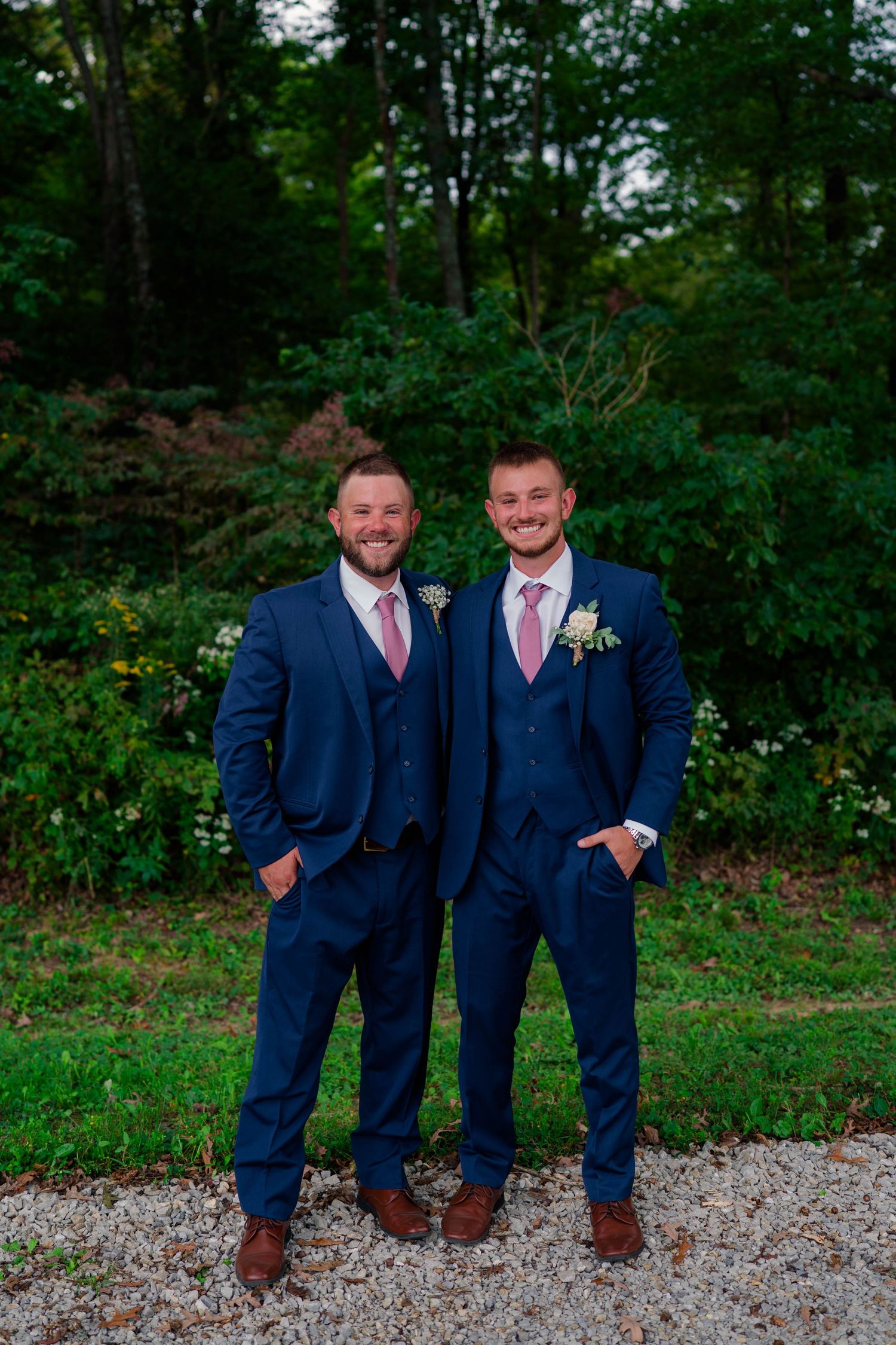 Two men in navy blue suits with pink ties pose outdoors at a wedding celebration.