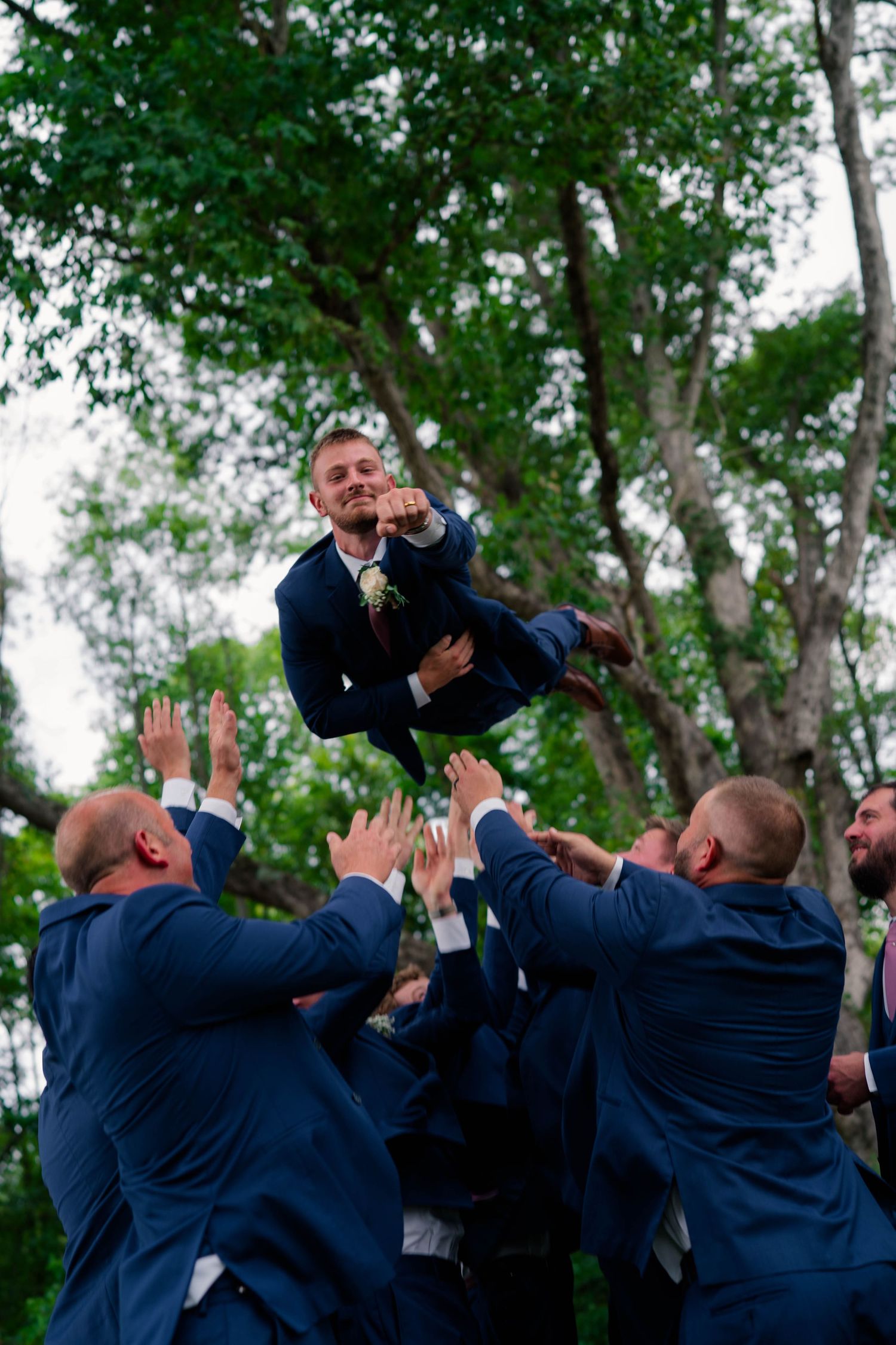 Groomsmen in navy suits tossing the groom into the air outdoors during a wedding celebration.