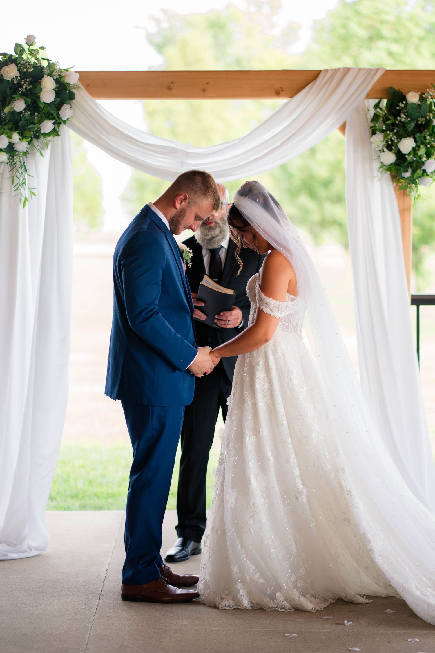 Bride and groom bow heads in prayer during outdoor wedding ceremony beneath elegant white draped arch with floral arrangements.