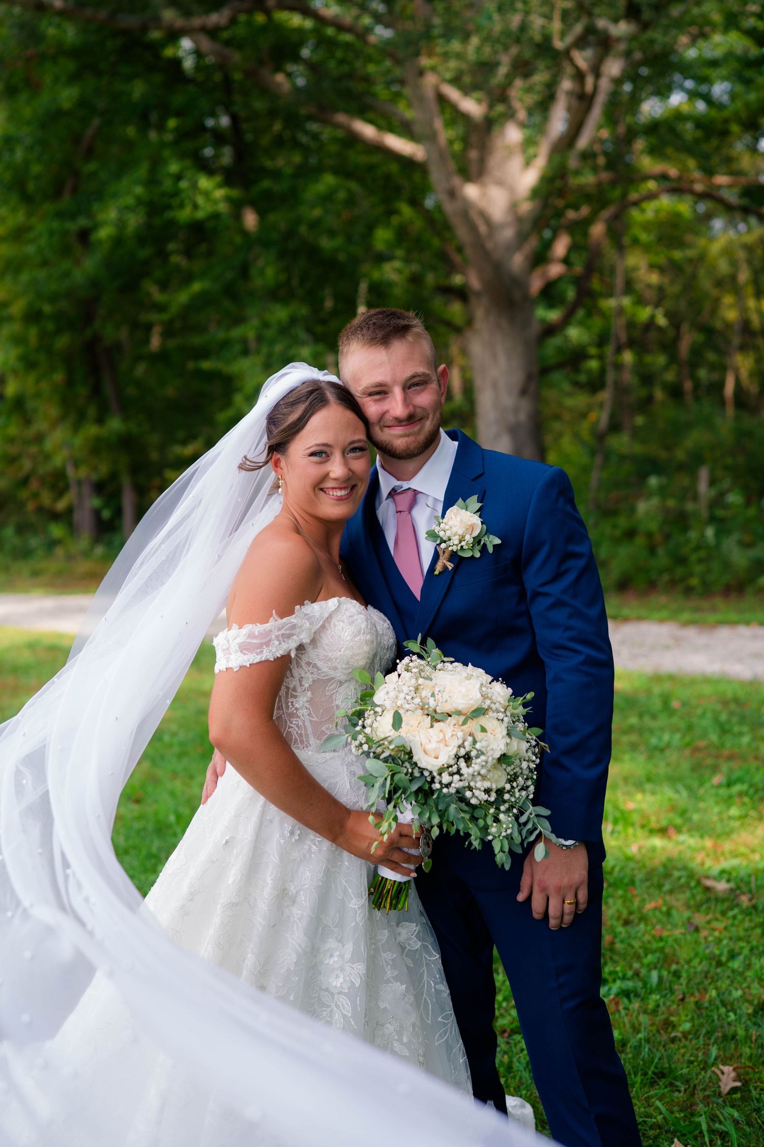 Smiling bride and groom pose outdoors in lush greenery, bride holding white bouquet with veil flowing in the breeze.