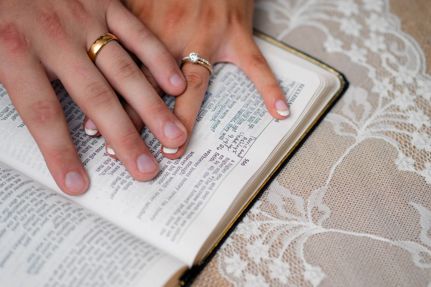 Bride and groom's hands with wedding rings rest together on open Bible on lace fabric.
