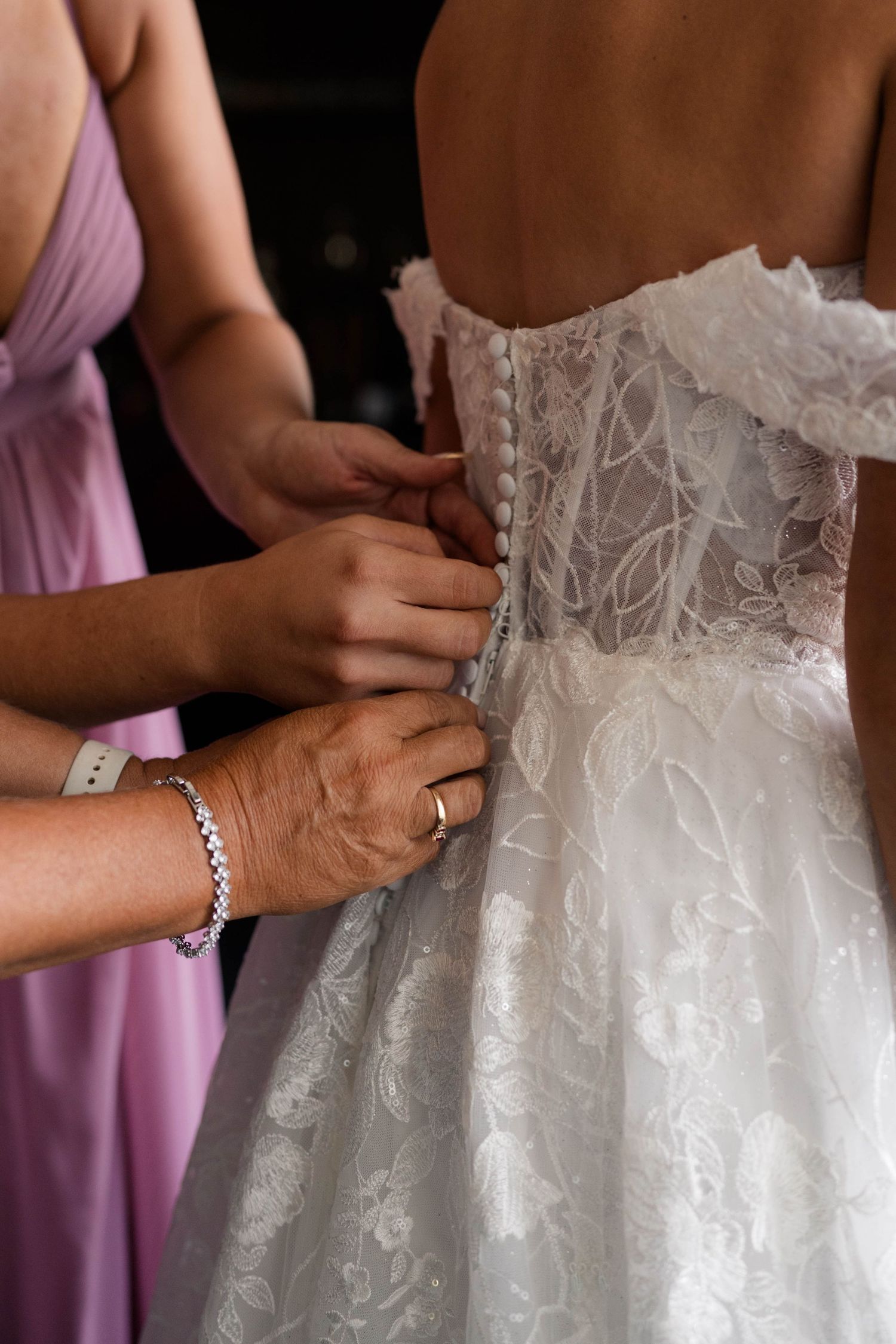 Bridesmaids helping bride button up her lace wedding dress from behind before the ceremony.