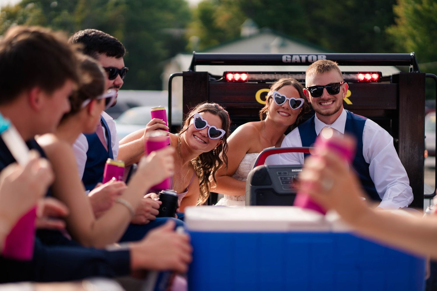 Bride and groom wearing heart sunglasses celebrate with wedding party on a Gator vehicle at outdoor reception.