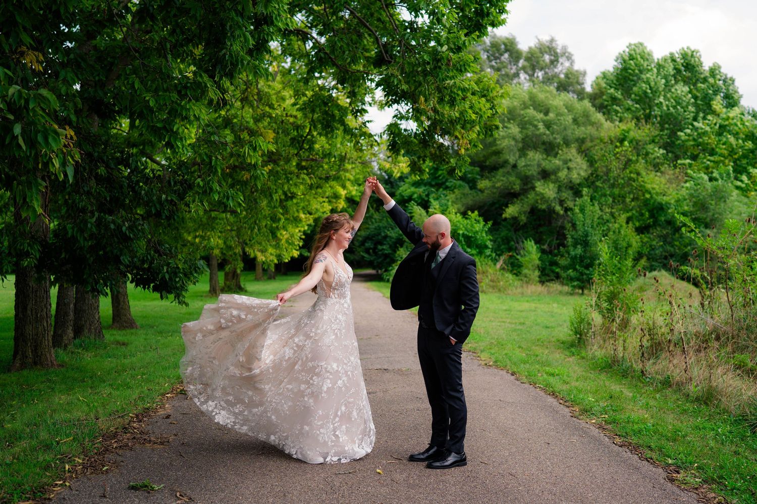 Groom twirling bride in flowing lace wedding gown on a tree-lined garden path during outdoor wedding portraits.