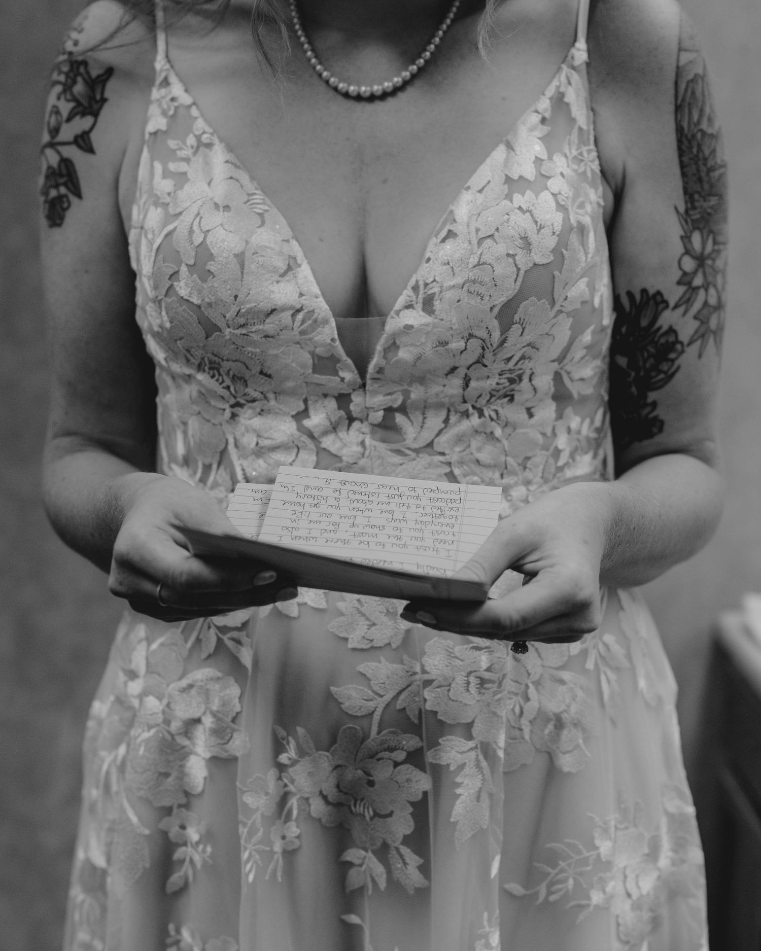 Bride in floral lace wedding gown reads handwritten vows while wearing pearl necklace and tattoos.