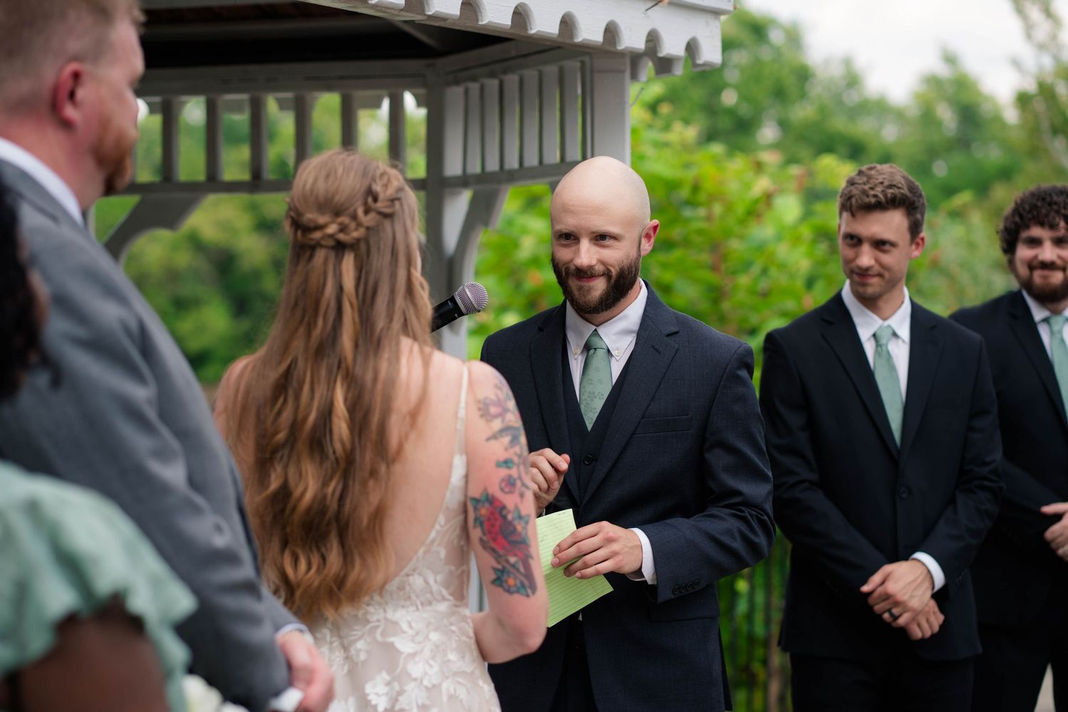 Bald groom smiling at bride while reading vows at outdoor wedding ceremony near gazebo.