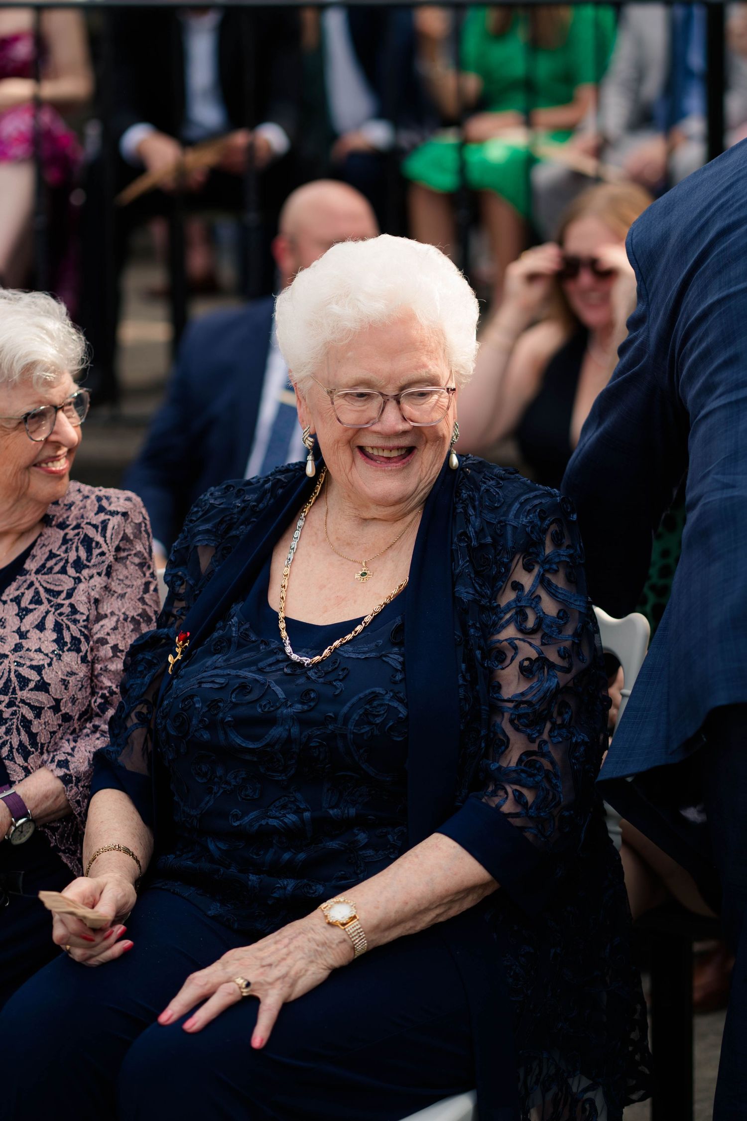 Elderly woman in navy lace dress smiles joyfully while seated at outdoor wedding ceremony.