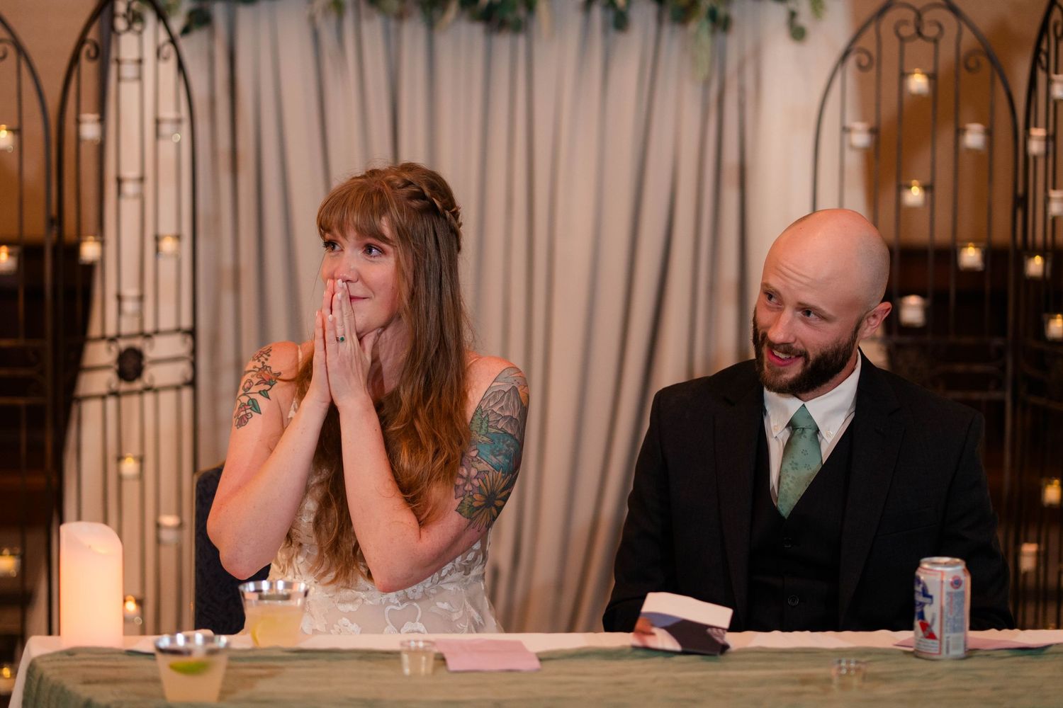 Bride covers her mouth in surprise while groom smiles during wedding reception speeches at decorated head table.