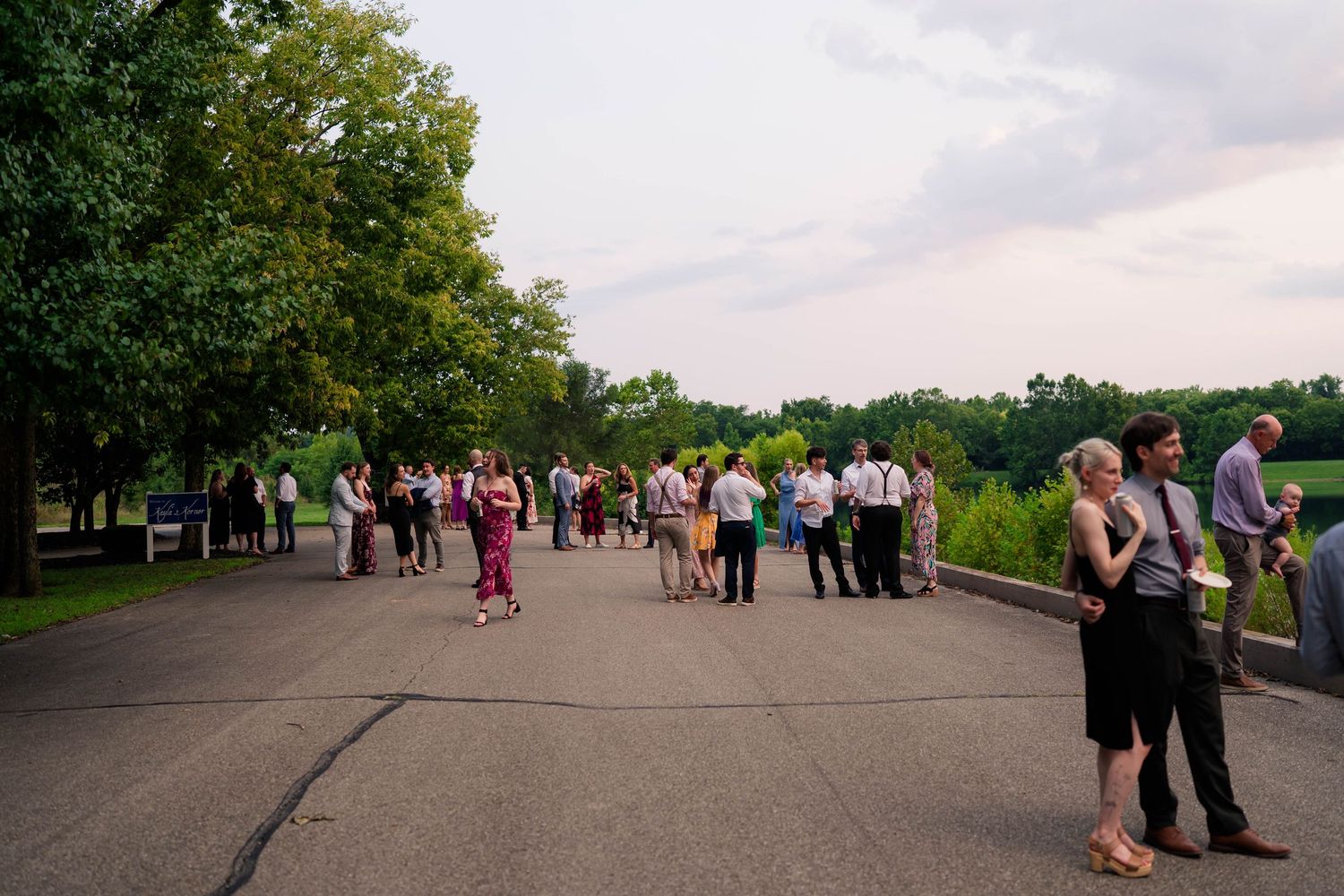 Wedding guests mingle outdoors on a paved area near a scenic lake at sunset.