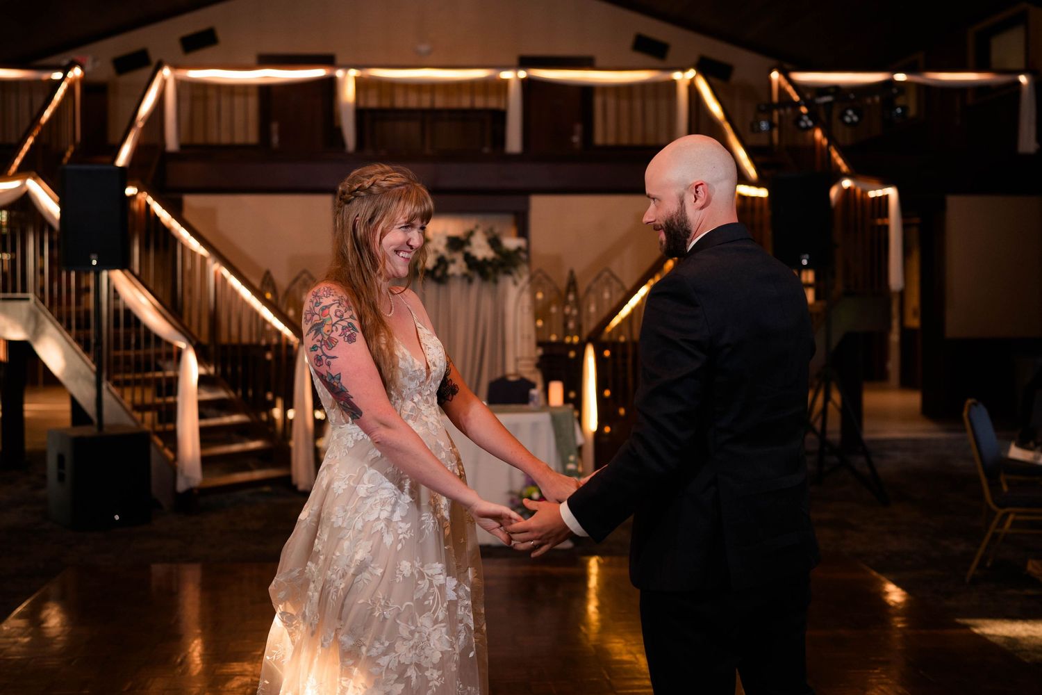 Bride and groom share a romantic first dance, holding hands and smiling in a warmly lit wedding reception venue.