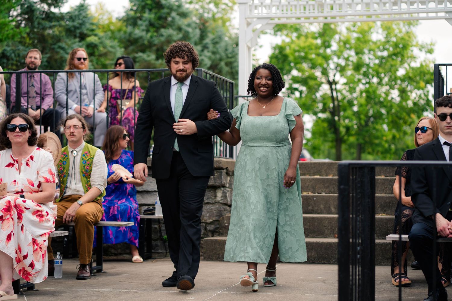 Groomsman in dark suit escorts a woman in a mint green dress down the wedding ceremony aisle outdoors.