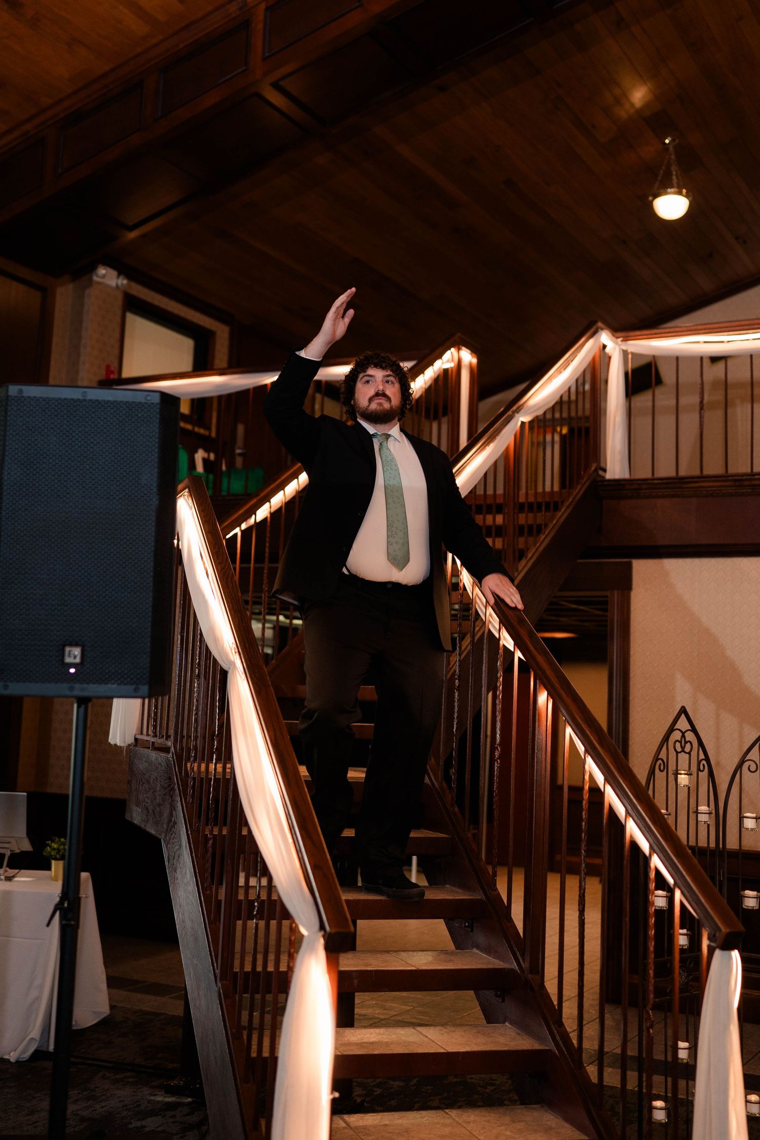 Groomsman in black suit dramatically poses on a wooden staircase at a wedding reception venue.