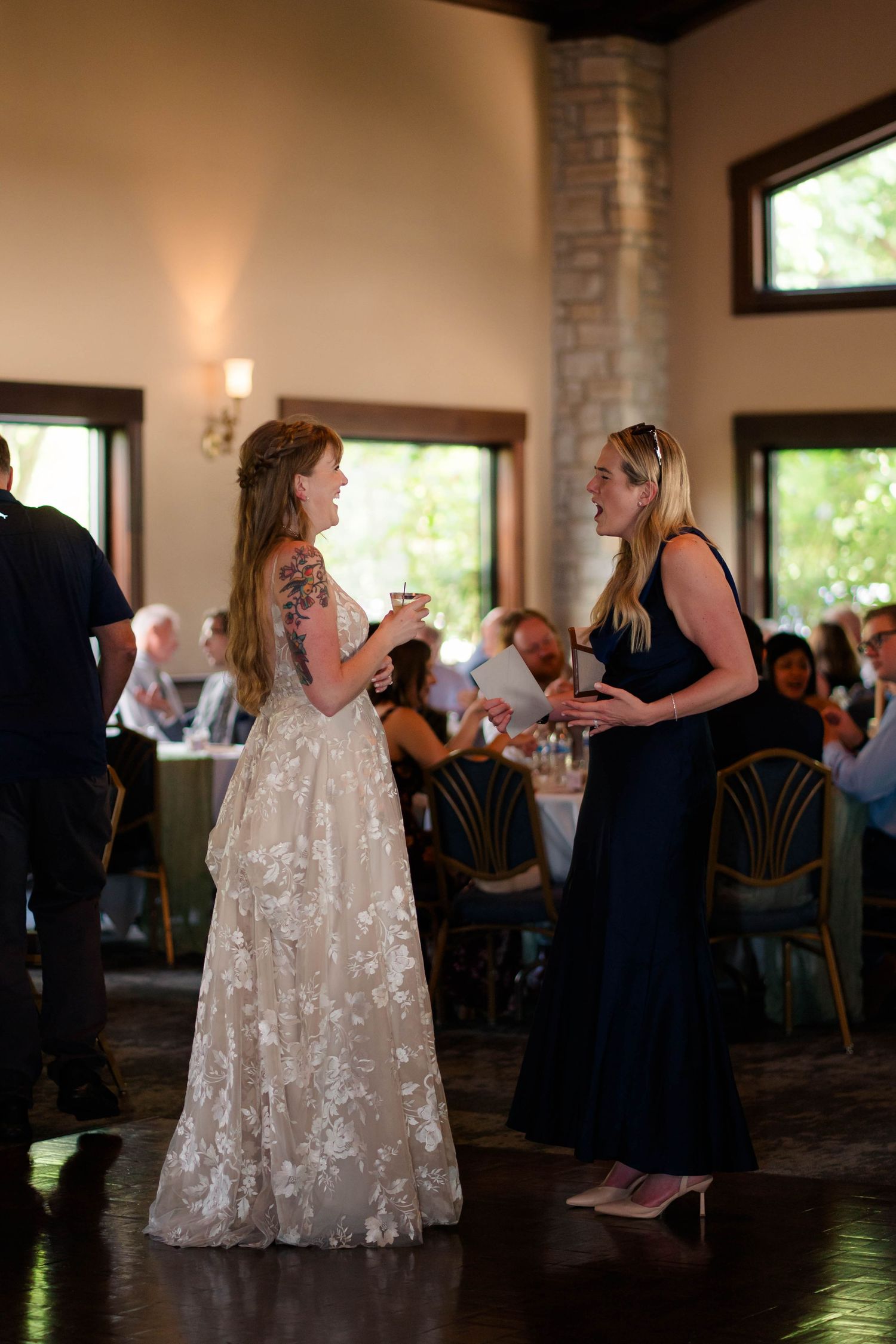 Bride in floral lace gown conversing with bridesmaid in navy dress at elegant wedding reception.