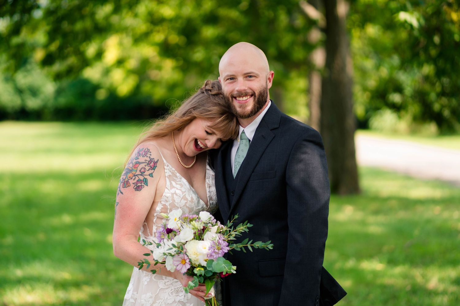 Smiling bald groom embraces tattooed bride holding bouquet in green park setting.