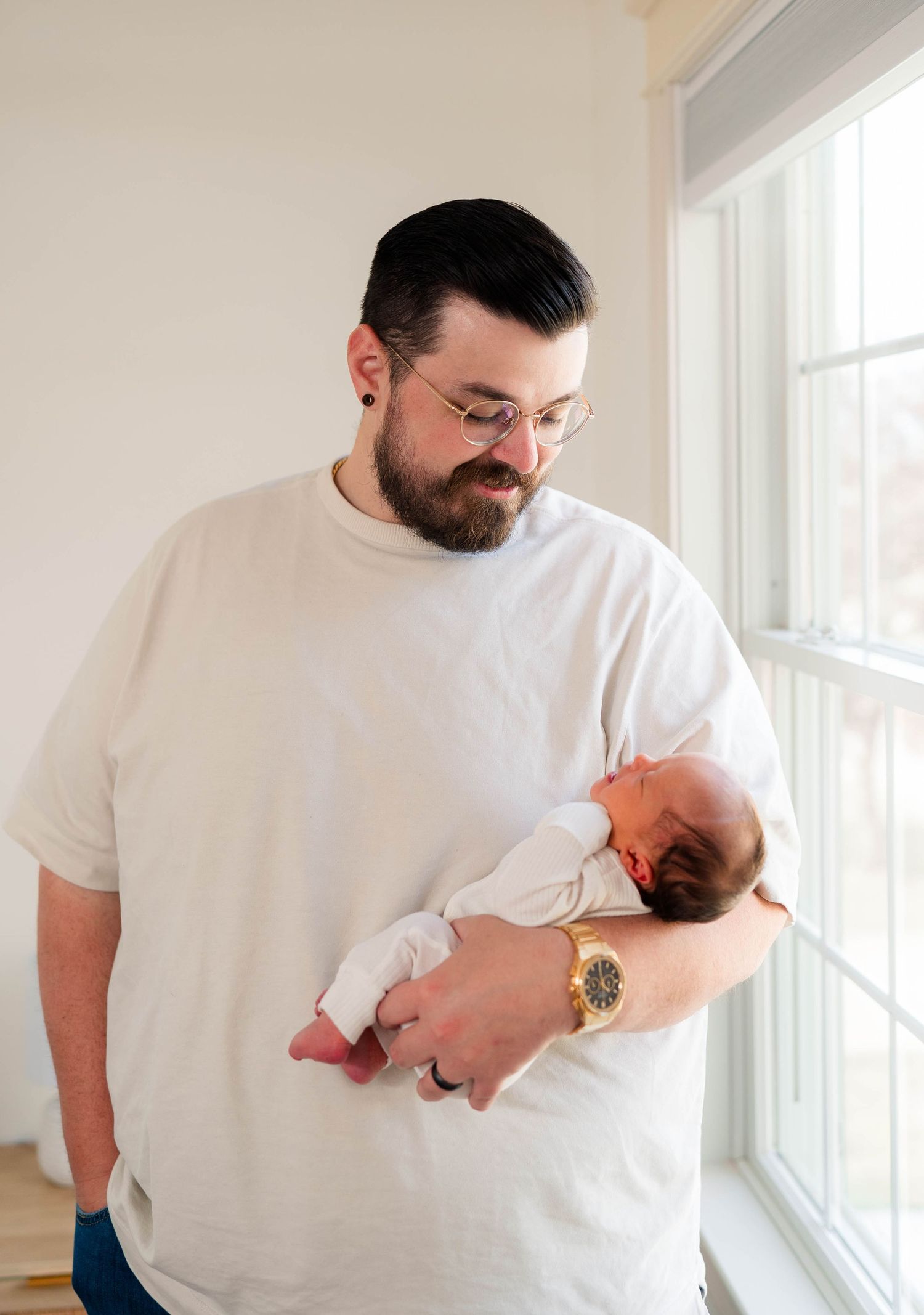 Bearded father in white t-shirt tenderly holds his newborn baby while standing by a bright window.