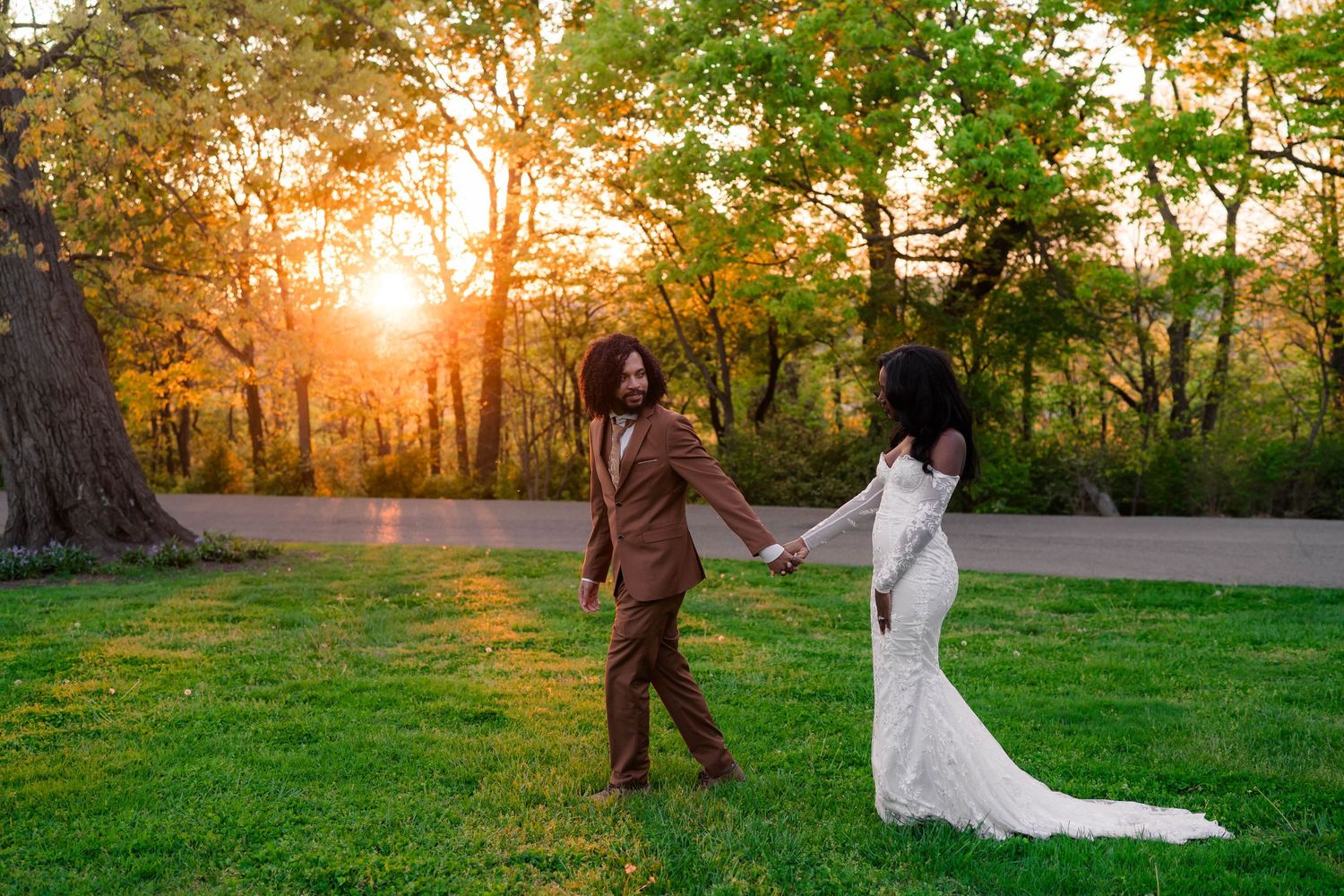Couple in wedding attire holding hands at sunset in a lush green park, bride in white gown and groom in brown suit.
