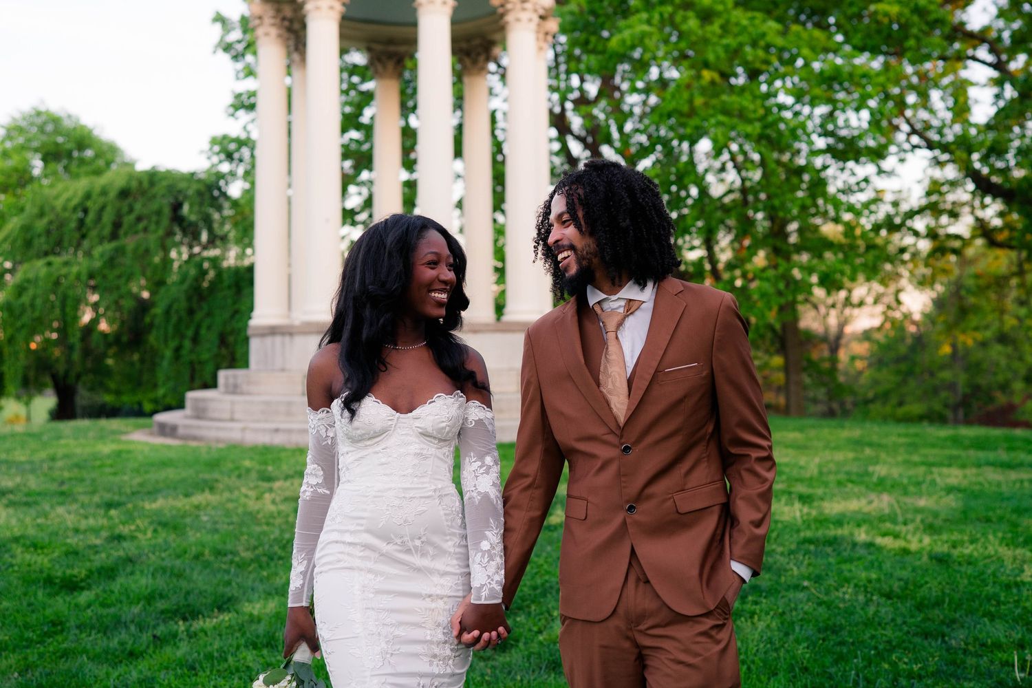 Joyful newlywed couple holding hands and laughing in front of white columns on lush green lawn.