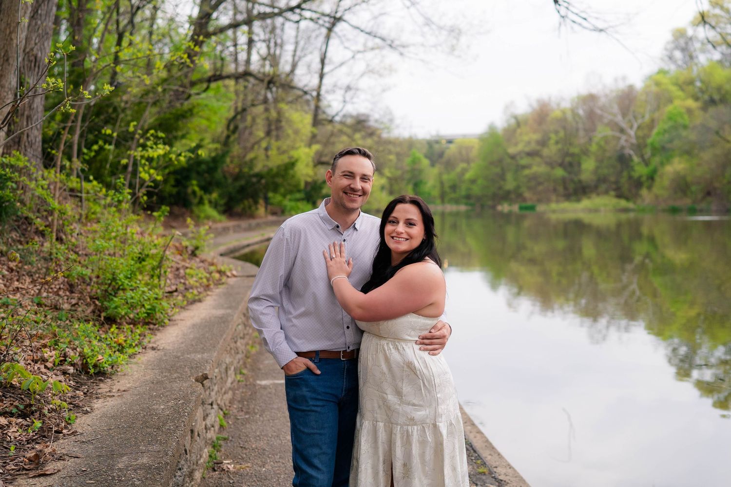 Couple smiling and embracing by a serene lake surrounded by lush green trees during an outdoor engagement session.