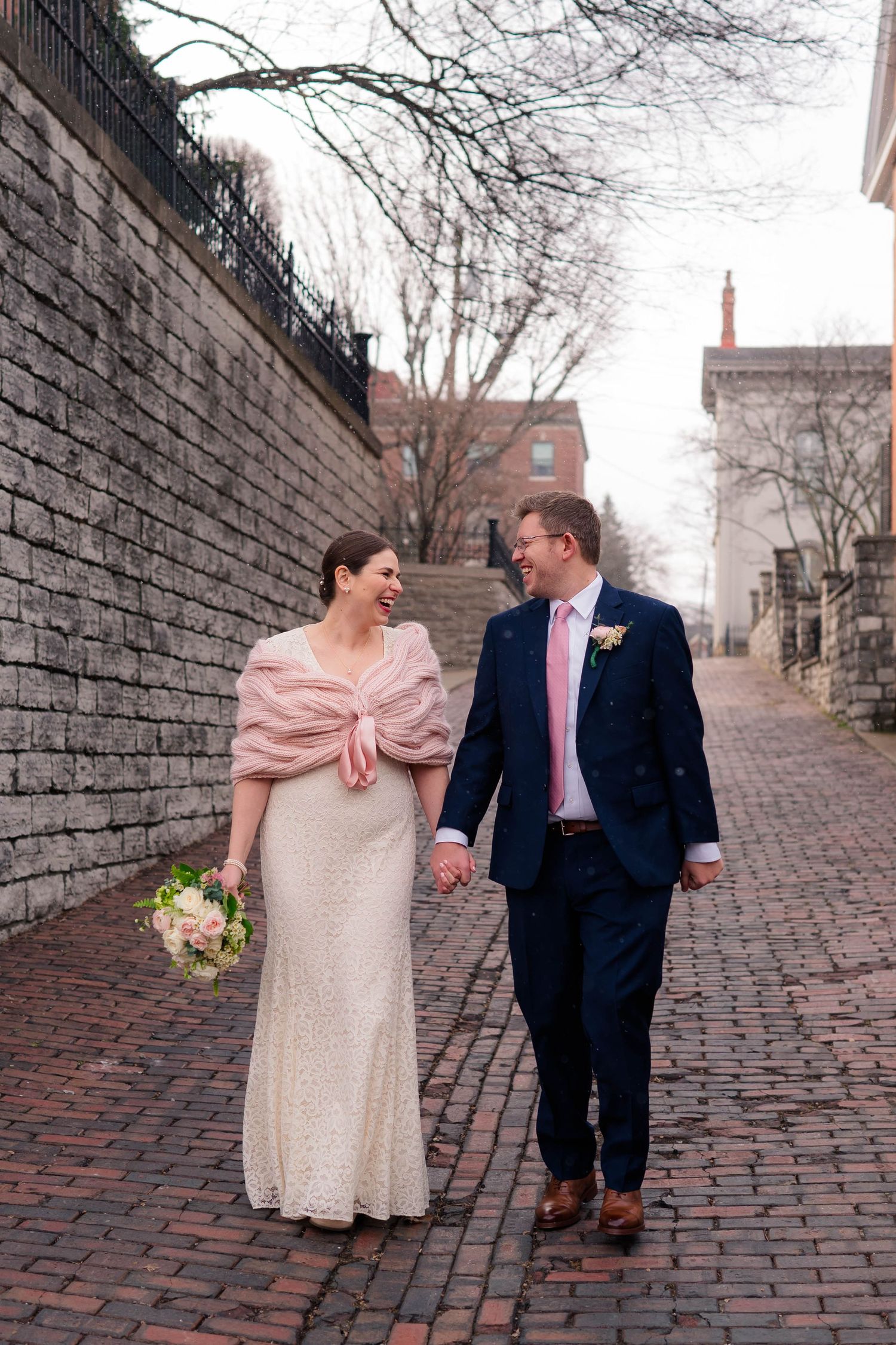 Bride and groom walk hand-in-hand on a brick alley, she in a lace gown with pink wrap, he in a navy suit.