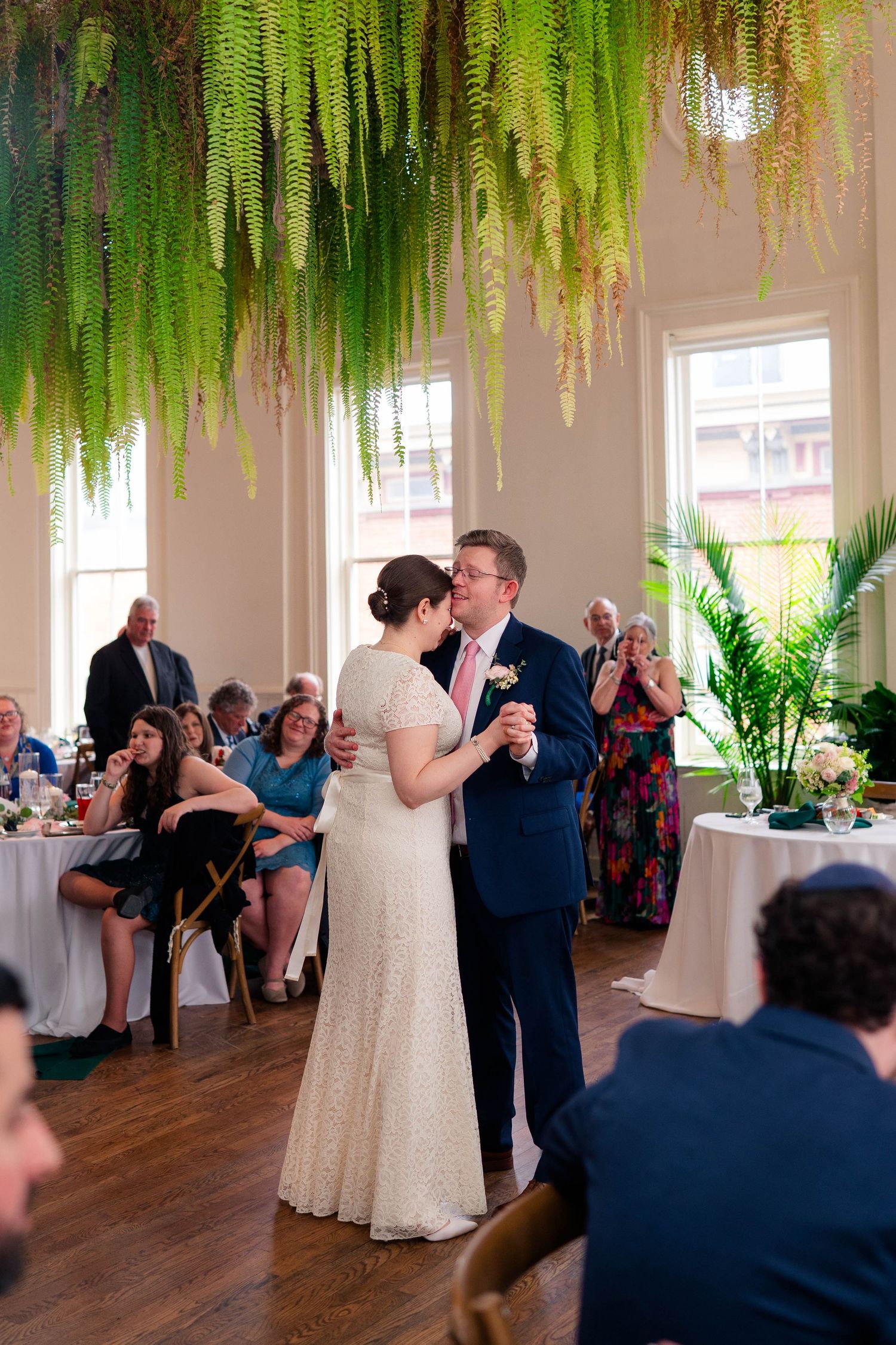 Couple sharing first dance under hanging ferns with guests applauding at wedding reception.